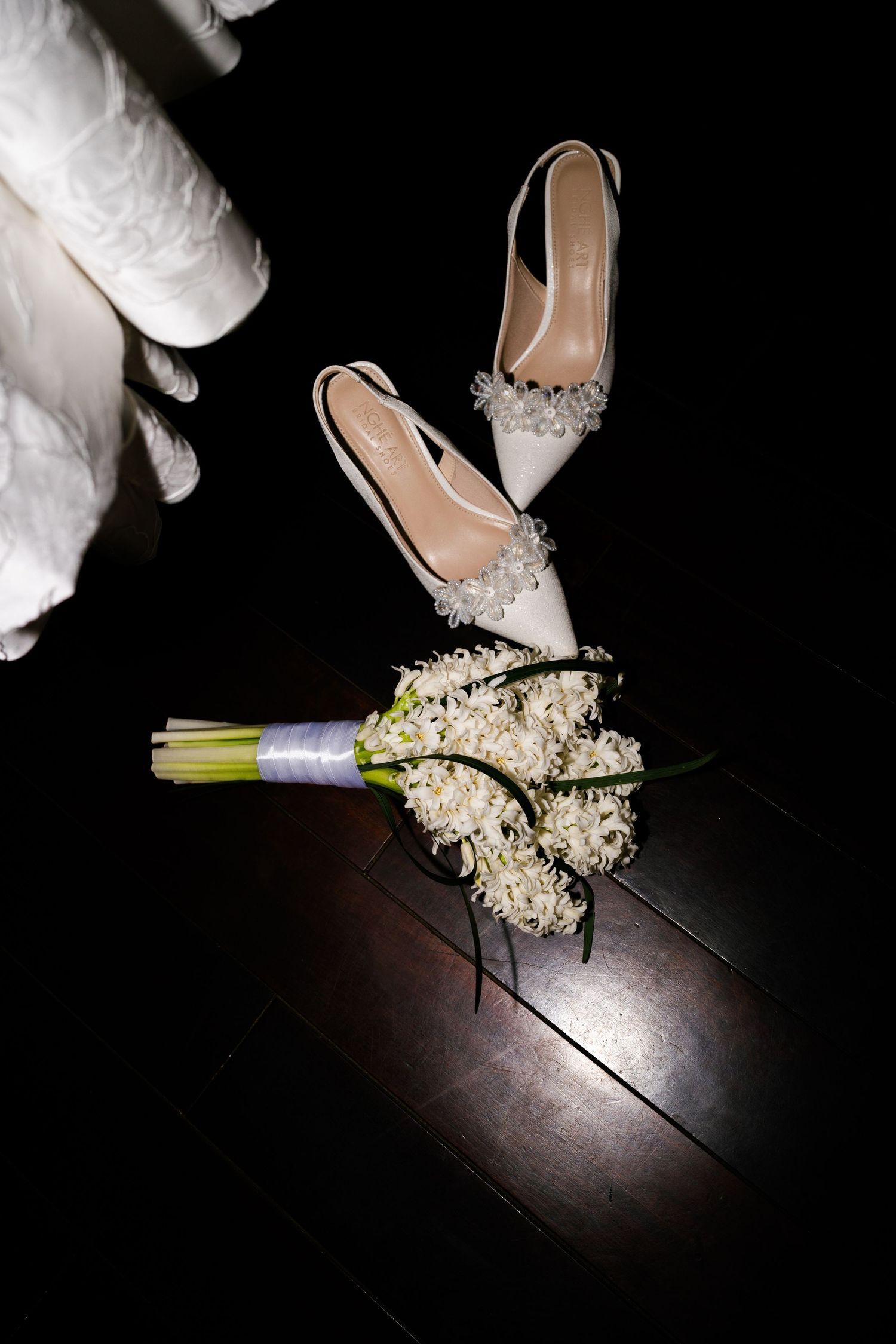 White bridal heels with crystal embellishments beside a white floral bouquet on dark floor.