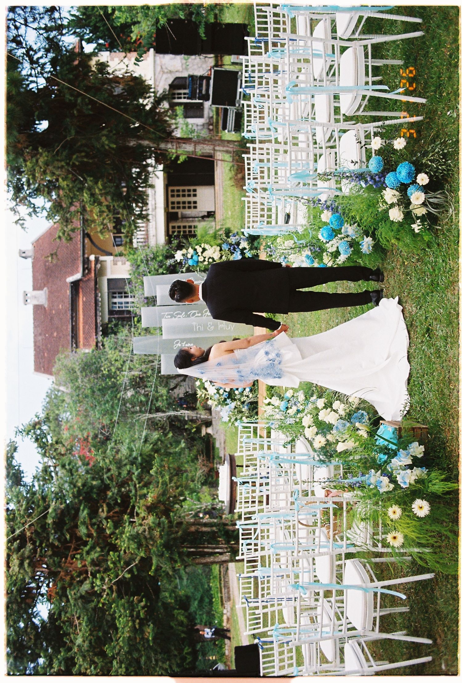 Bride and groom standing at outdoor wedding ceremony aisle lined with white chairs and blue floral arrangements.