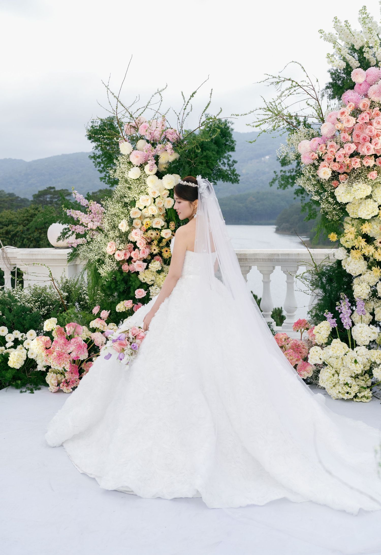 Bride in a white ballgown and veil stands before a lush floral arch with pink and yellow blooms overlooking a lake.