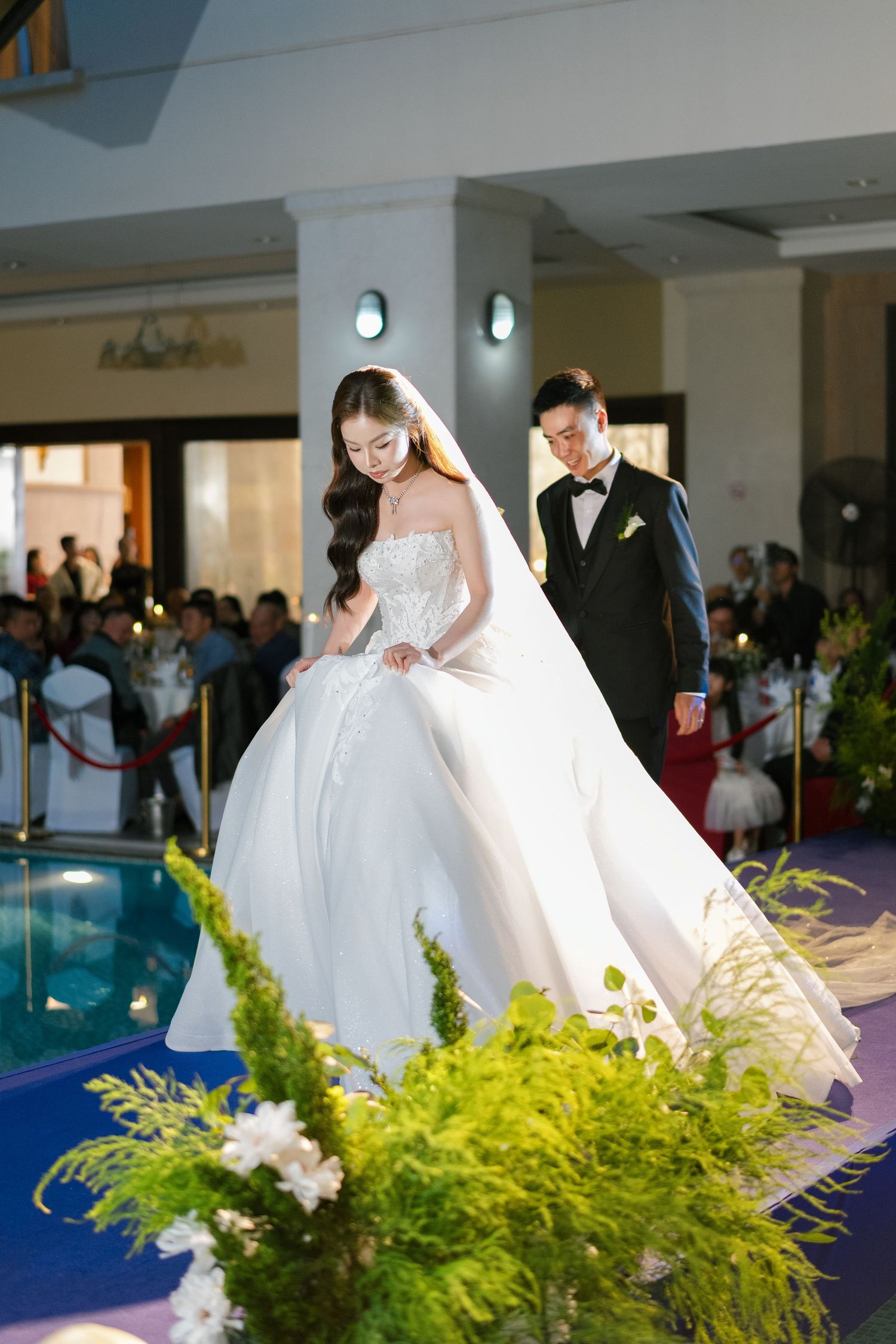 Bride in white ballgown and groom in black tuxedo walk down aisle at elegant wedding ceremony venue.