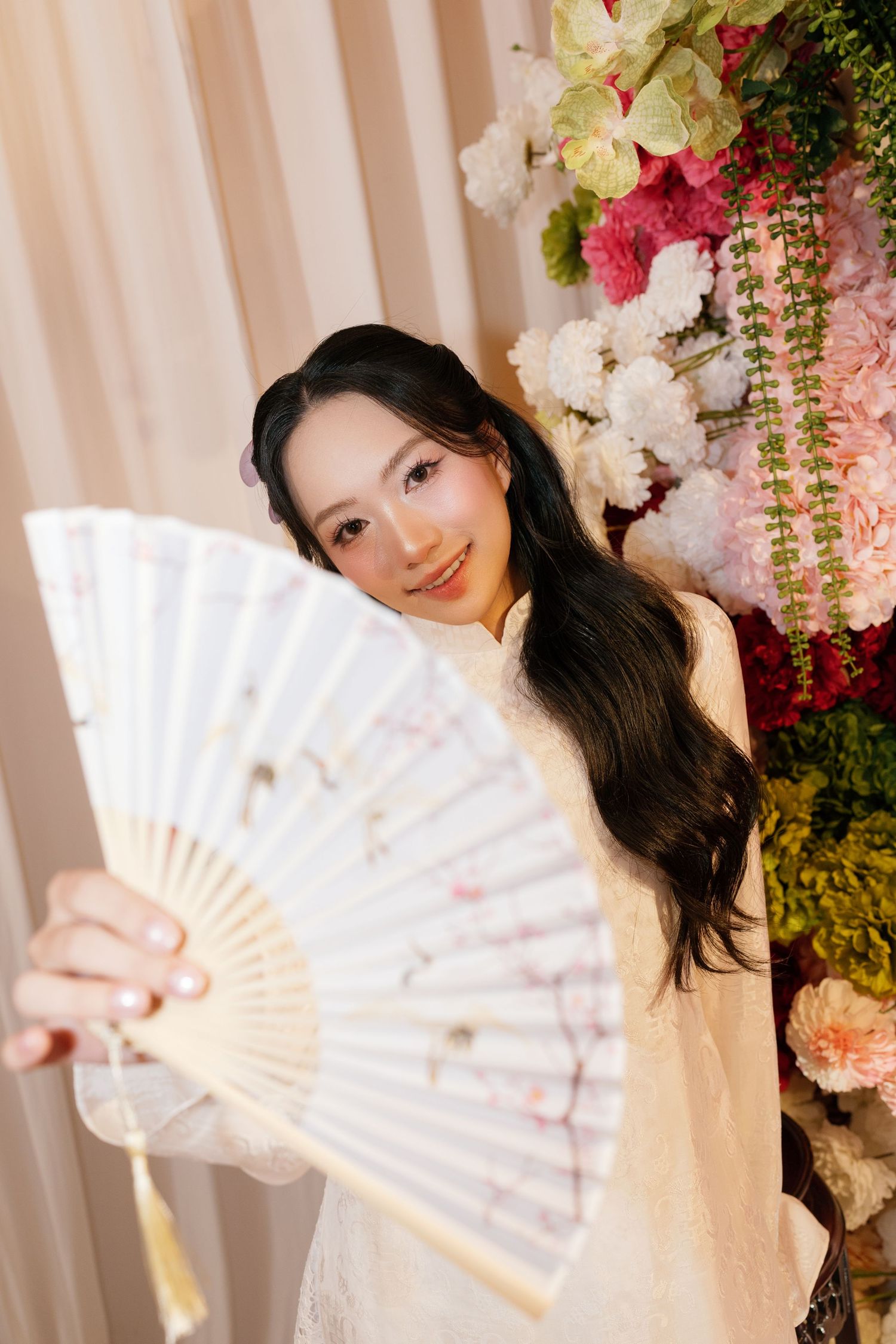 Young Asian woman in white dress holds decorative floral fan near pink and white flower arrangement.