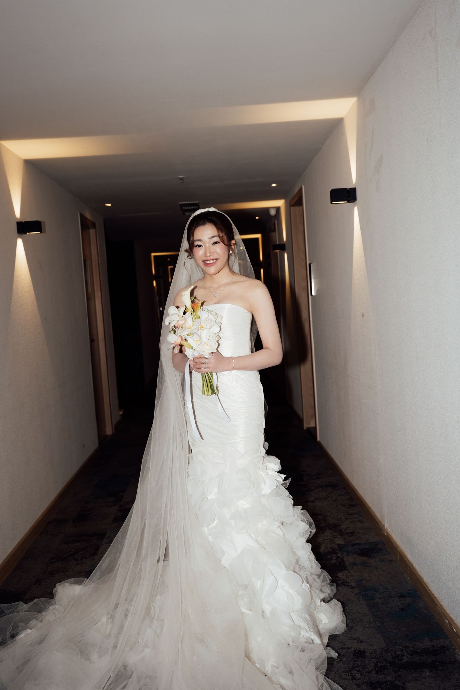 A smiling bride in a strapless mermaid wedding dress with veil holds a bouquet while standing in a hotel corridor.
