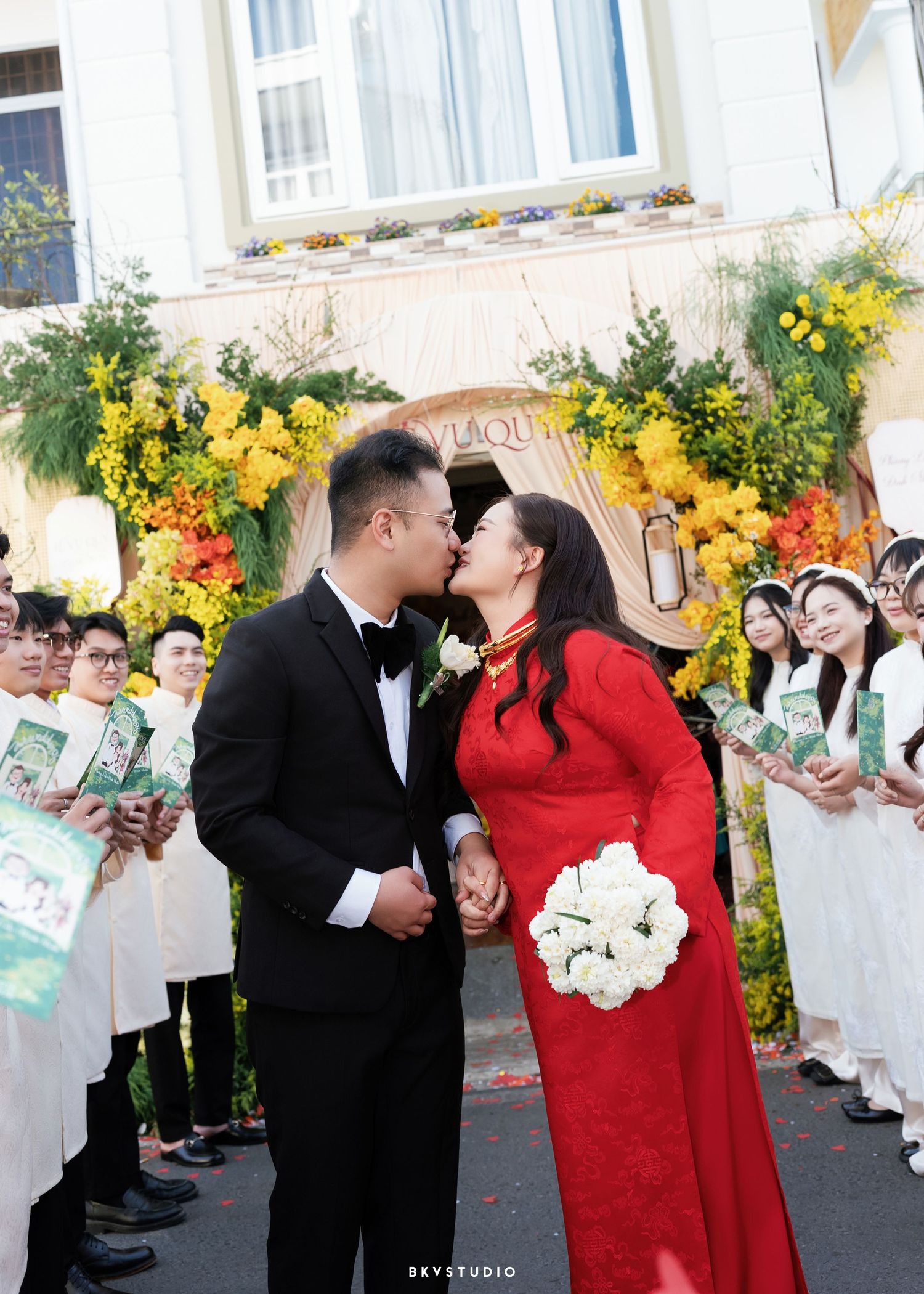 Vietnamese couple kissing at traditional wedding, bride in red ao dai holding white bouquet, groom in black tuxedo.
