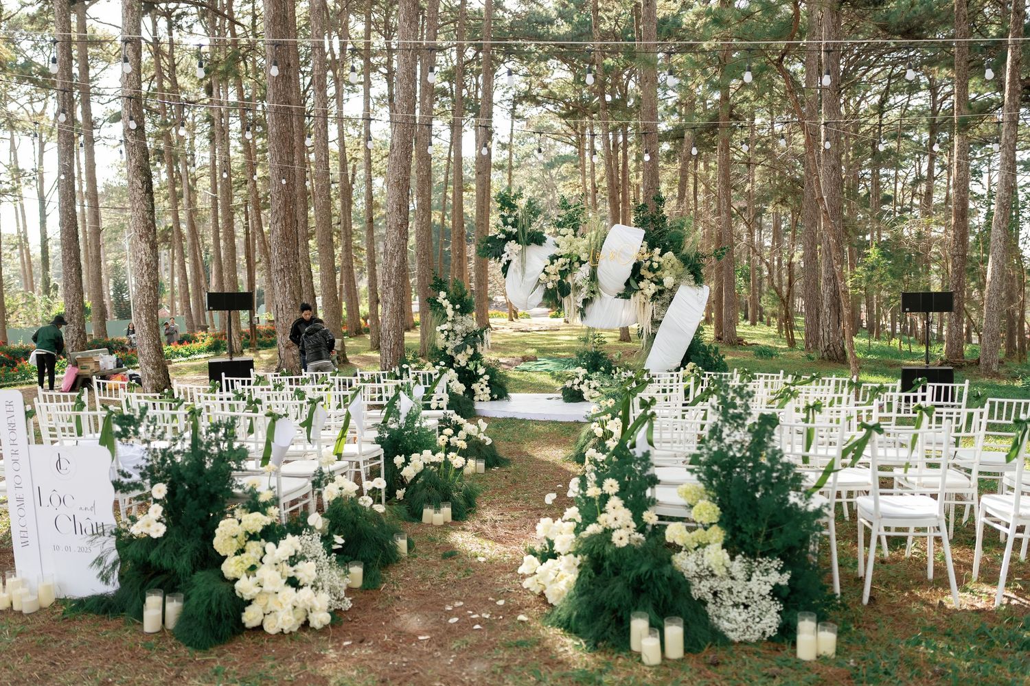 Outdoor forest wedding venue with white chairs, candles, and white floral arrangements lining the aisle.
