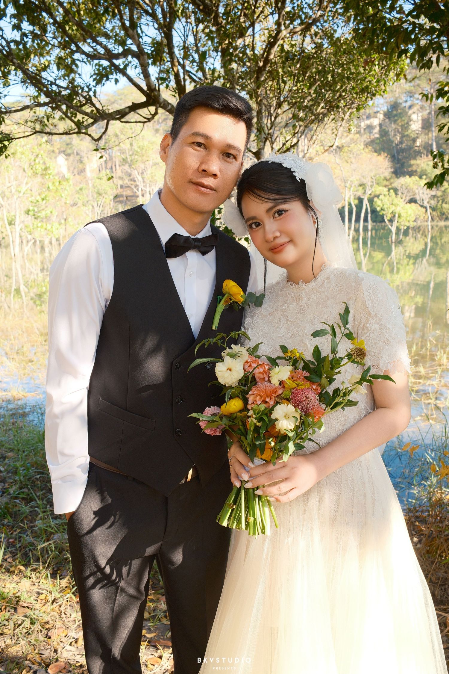 Bride and groom smile together holding a vibrant bouquet near a reflective lake with autumn foliage.