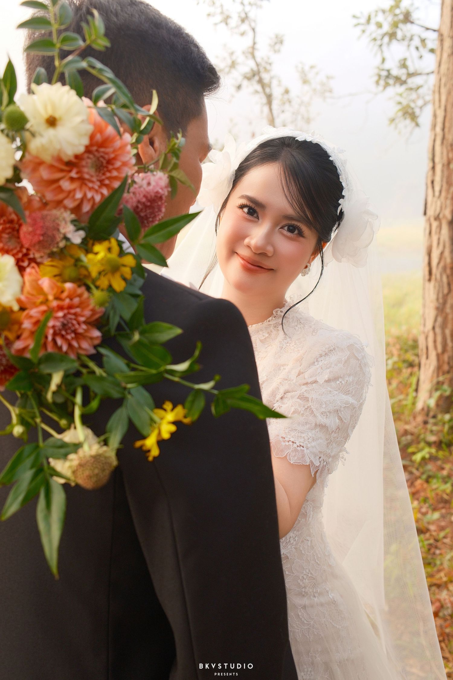Bride in lace wedding dress and veil smiles over groom's shoulder beside colorful floral bouquet outdoors.