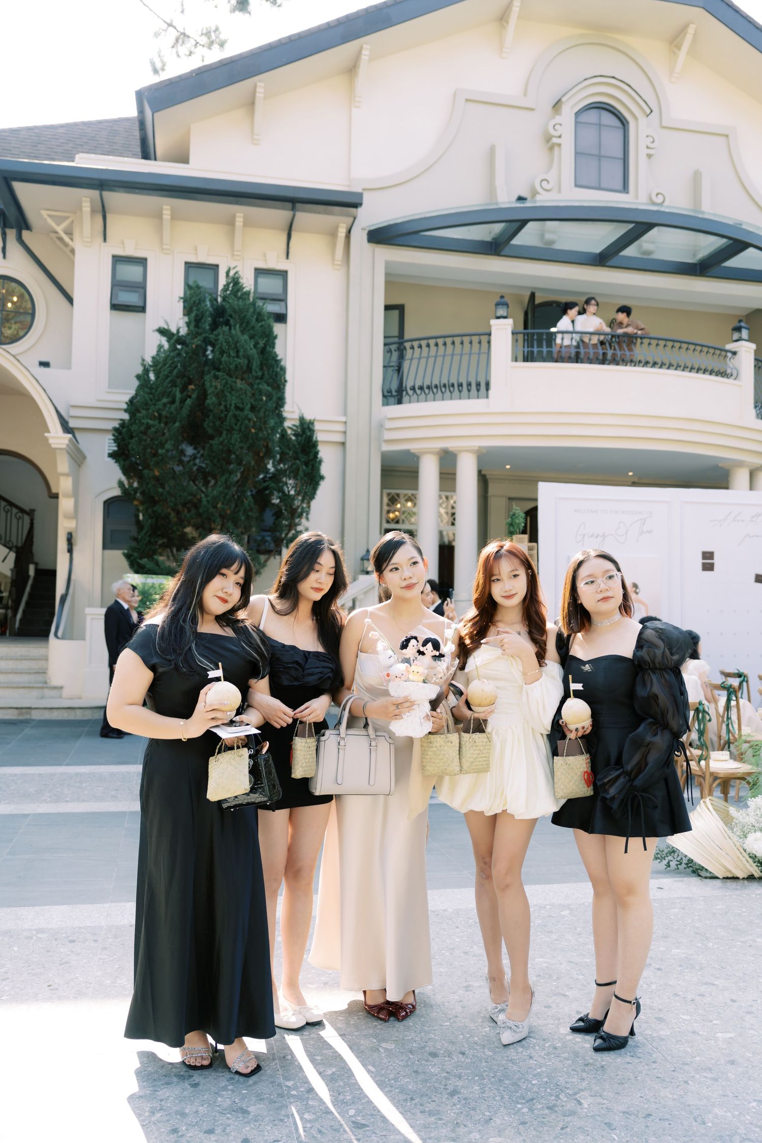 Group of five women in black and nude dresses pose outside grand wedding venue.