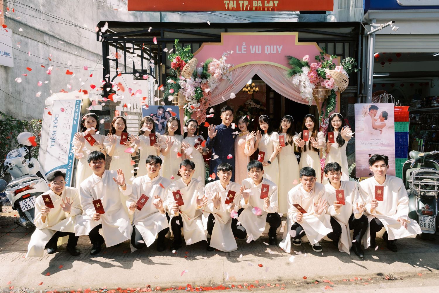 Large Vietnamese wedding party in traditional white ao dai poses outside decorated venue entrance.
