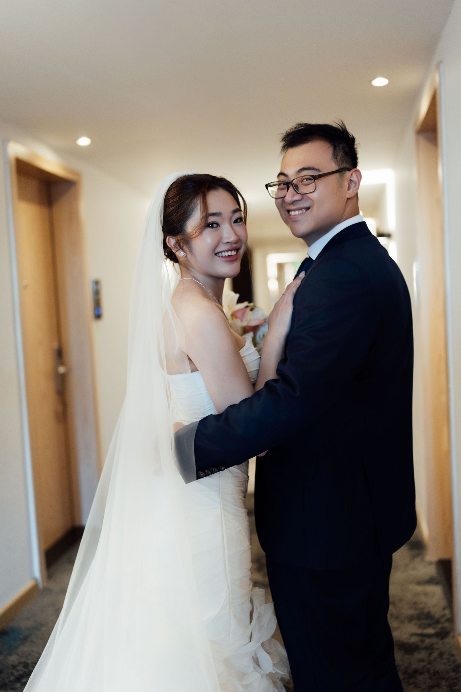 Smiling bride in white gown and veil with groom in tuxedo posing together in hotel hallway on wedding day.