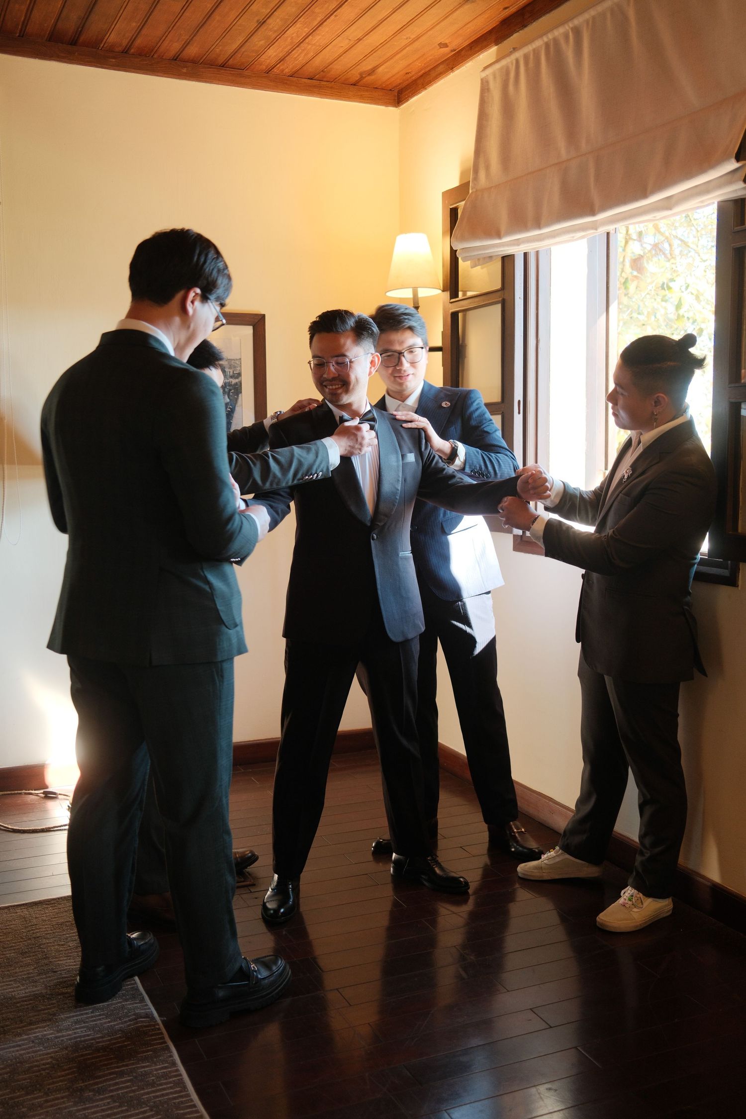 Groomsmen help the groom adjust his bow tie and suit in a warmly lit wedding preparation room.