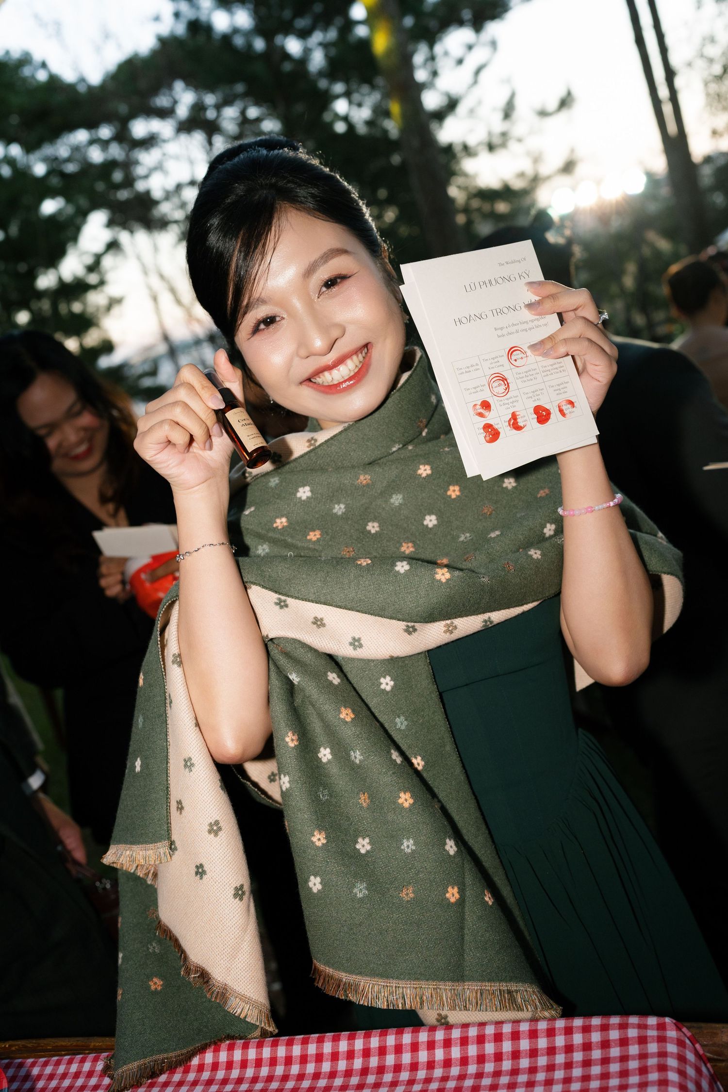 Smiling wedding guest in green dress and floral scarf holding up a wedding program card outdoors.