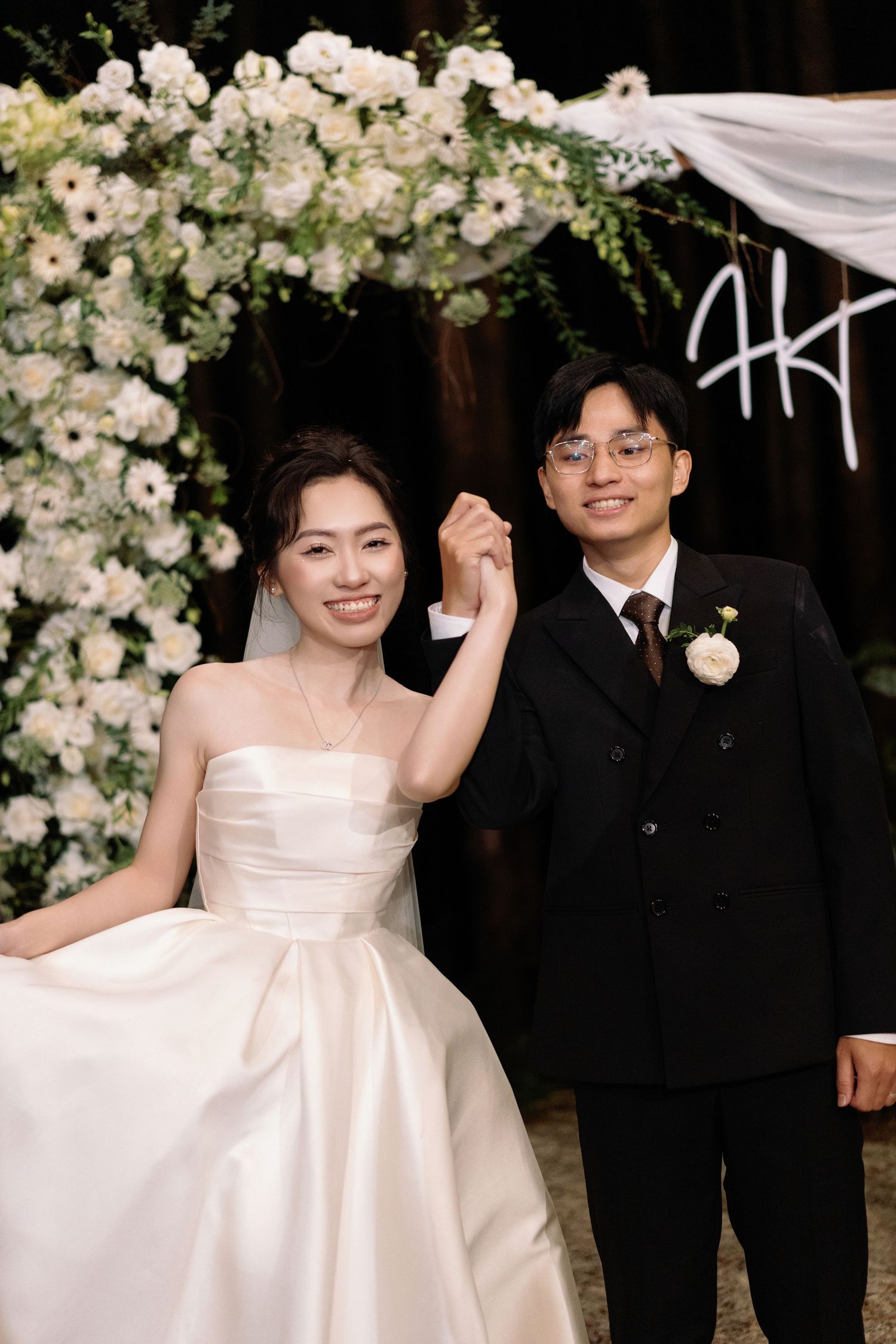 Asian bride and groom hold hands and smile before floral arch backdrop at elegant wedding reception.