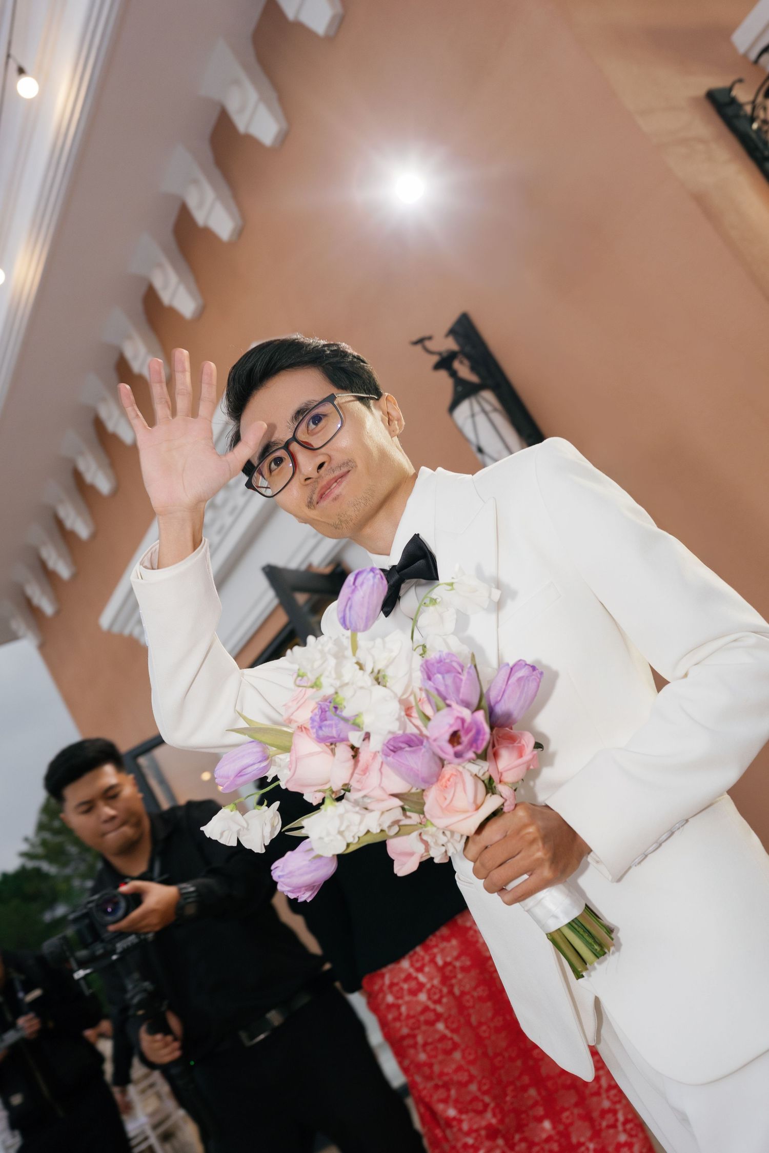 Groom in white tuxedo holding purple and pink bouquet, waving at wedding ceremony outdoors.