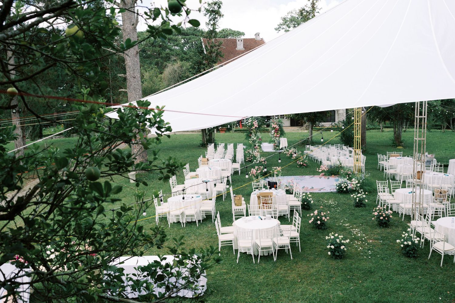 Elegant outdoor wedding reception under white tent with round tables, white chairs, and floral arrangements on green lawn.
