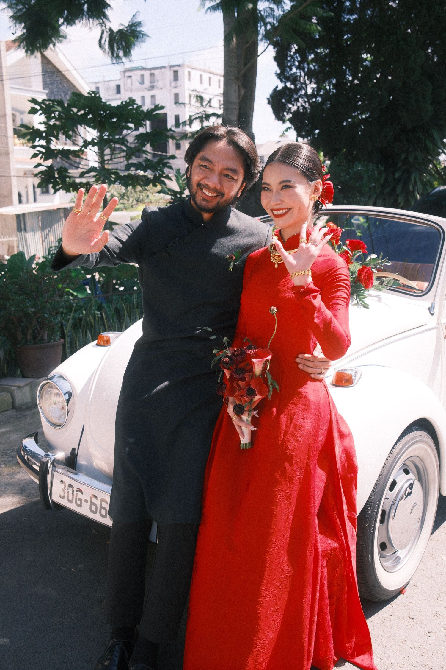 Couple in traditional Vietnamese wedding attire pose by a white vintage car, woman in red ao dai, man in black.