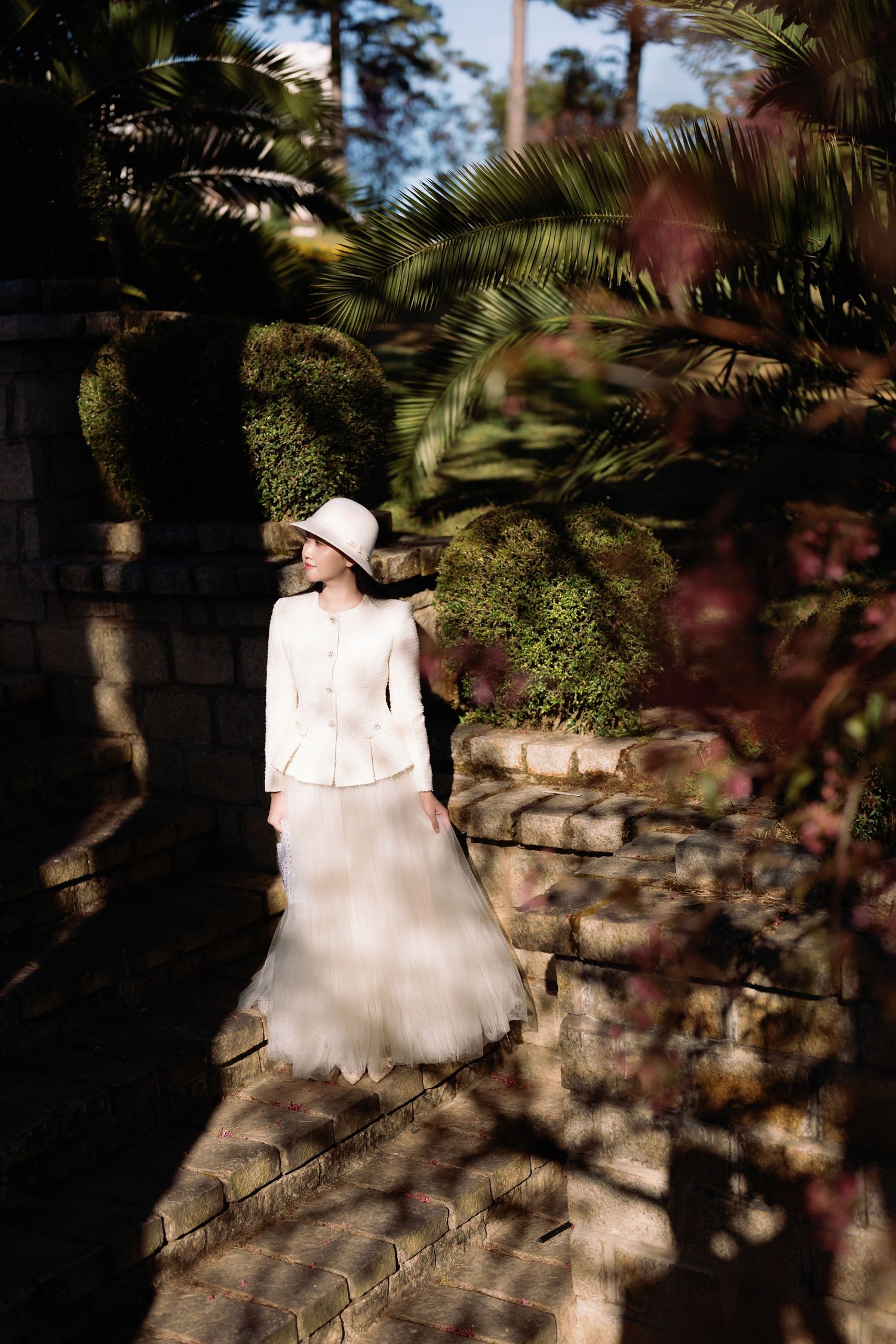 A bride in a white jacket and tulle skirt poses in a sunlit garden surrounded by lush greenery and stone walls.