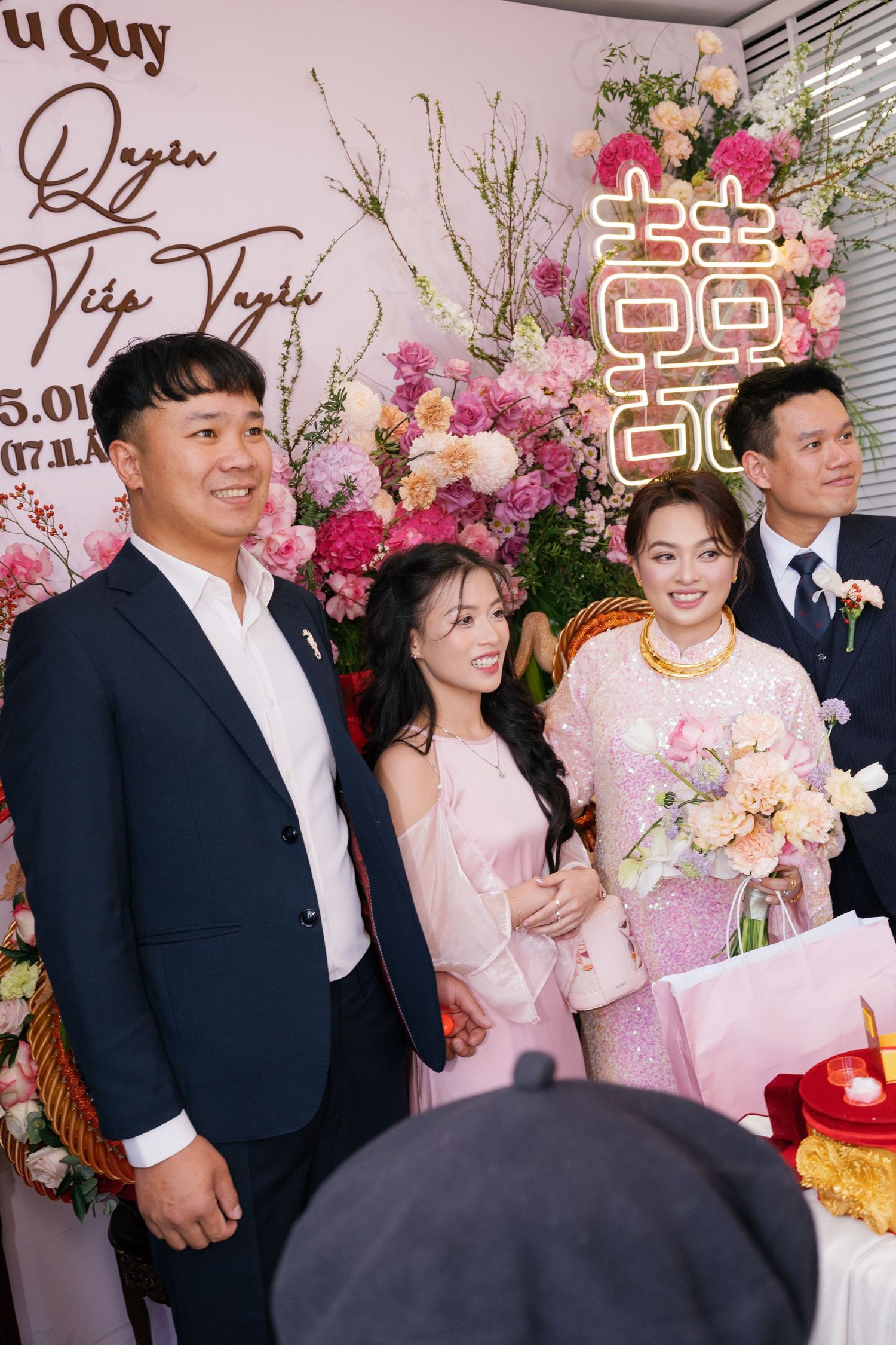 Vietnamese wedding party poses in pink ao dai and suits before floral double happiness backdrop.