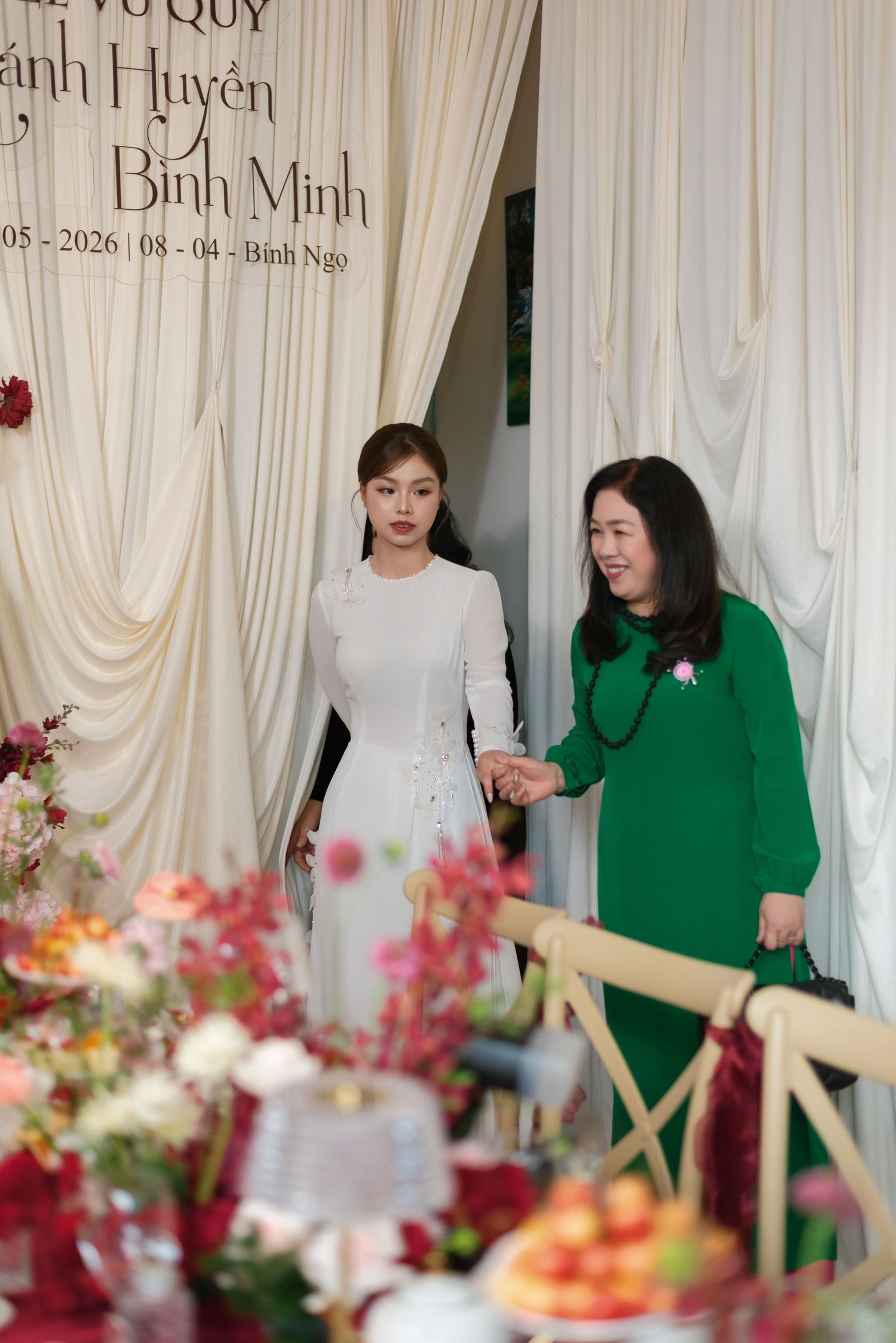 Two women in elegant attire, one in white ao dai and one in green dress, at a Vietnamese wedding ceremony.