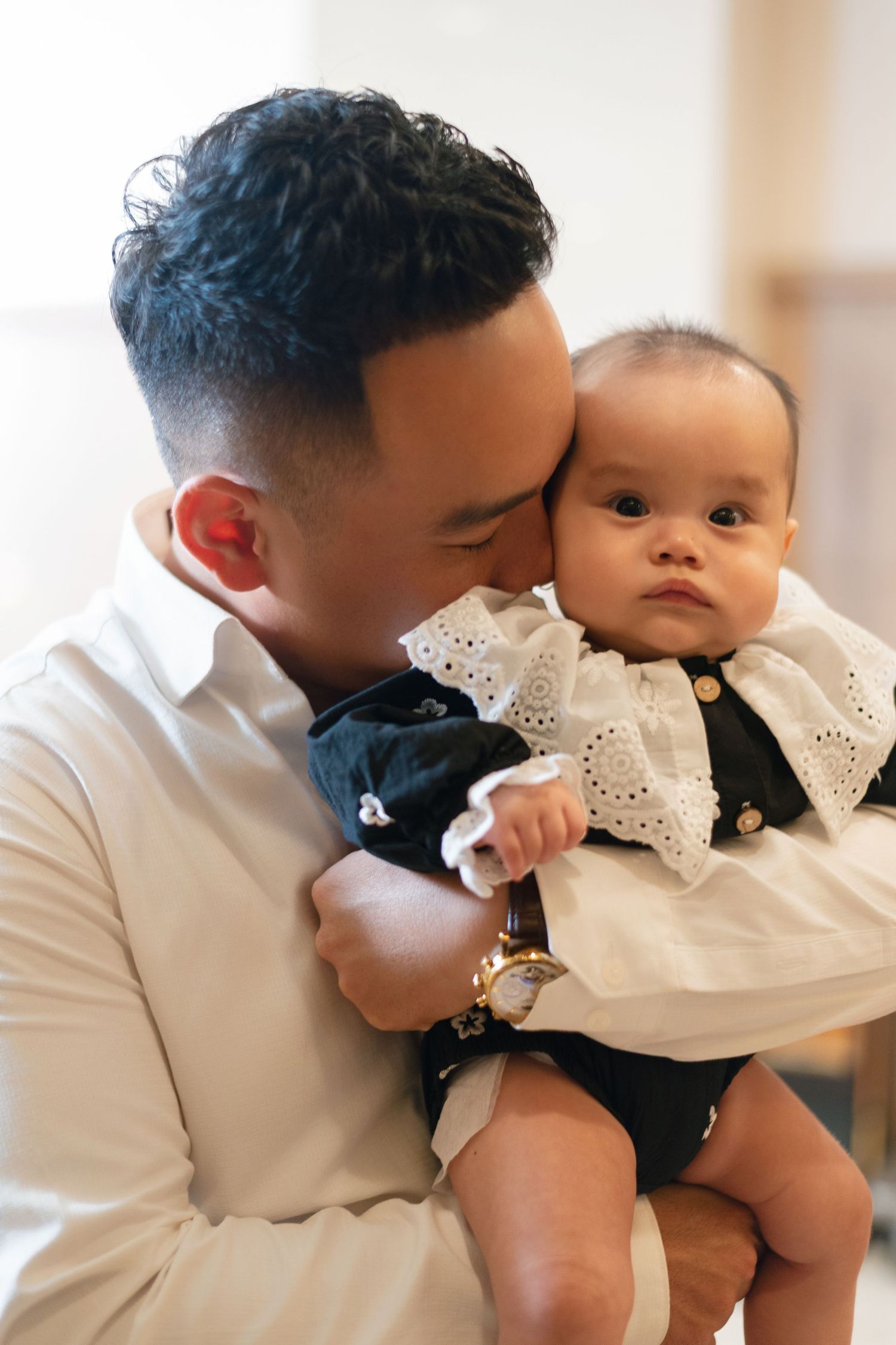 A father tenderly kisses his baby dressed in a dark outfit with white lace collar while holding them close.