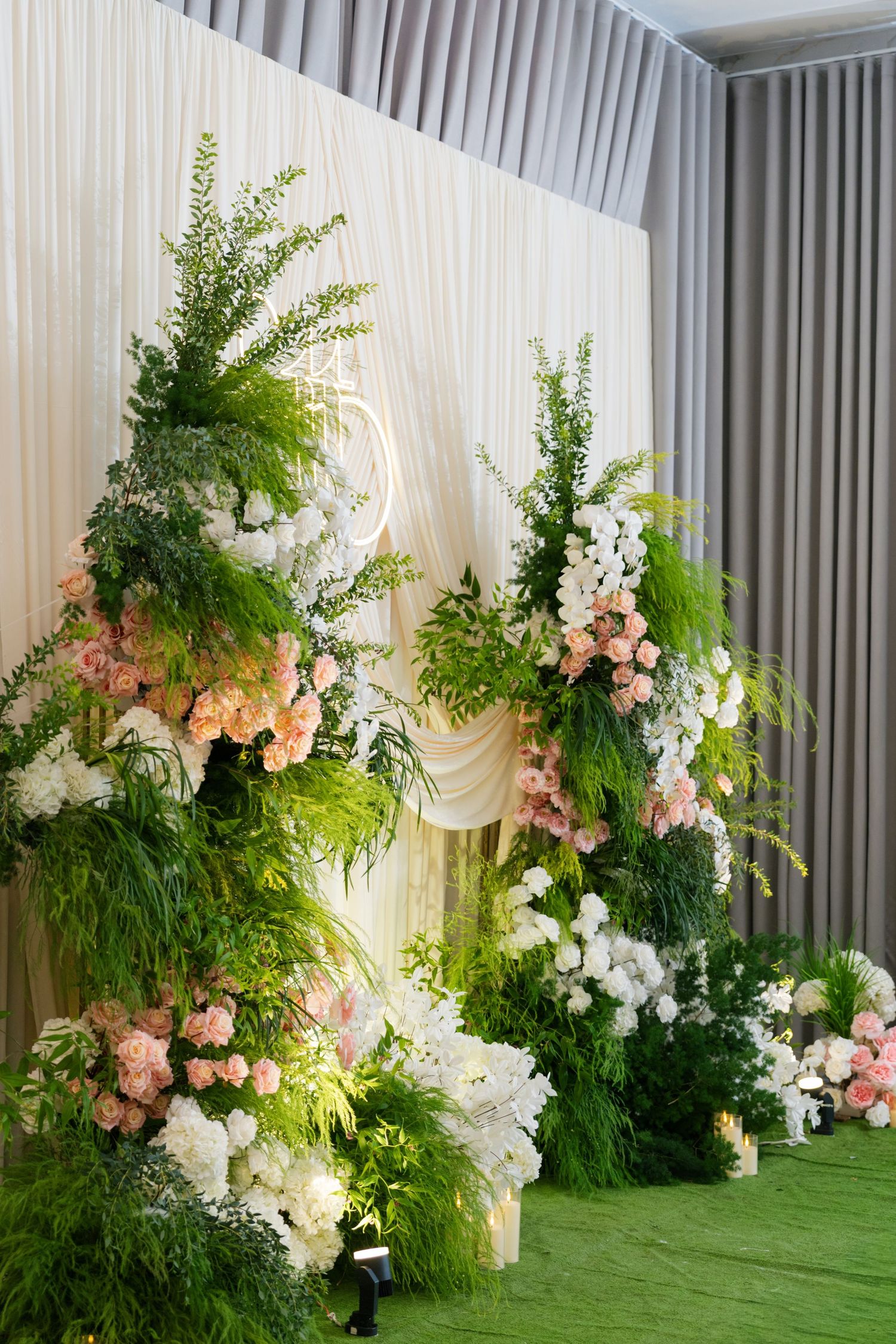Elegant wedding backdrop with lush green ferns, white draping, and pink and white floral arrangements on green carpet.