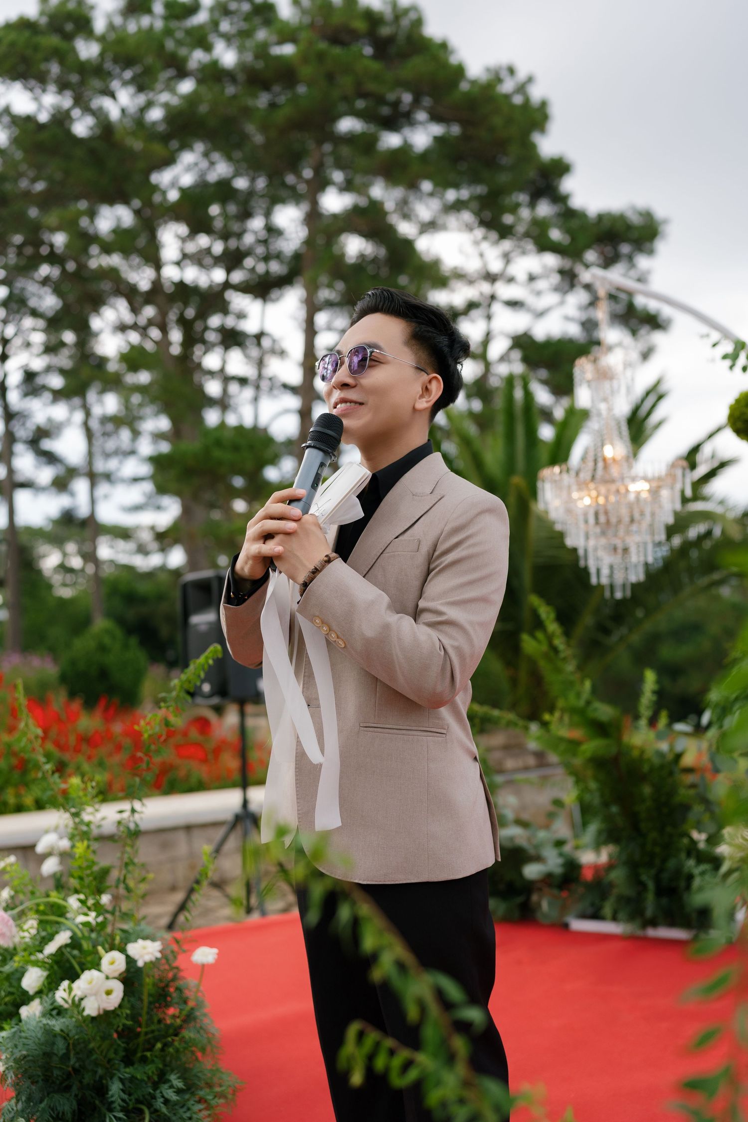 A stylish emcee in a beige blazer and sunglasses speaks into a microphone at an outdoor wedding ceremony.