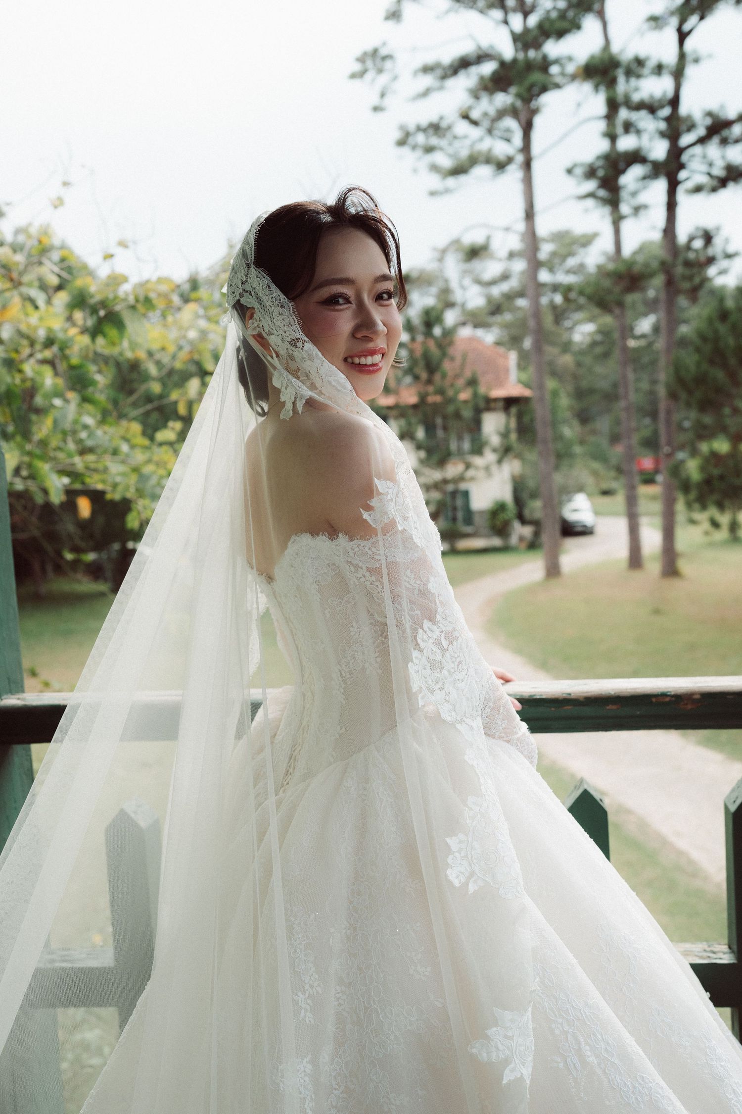 Smiling Asian bride in white lace wedding gown and veil stands on balcony surrounded by lush greenery.