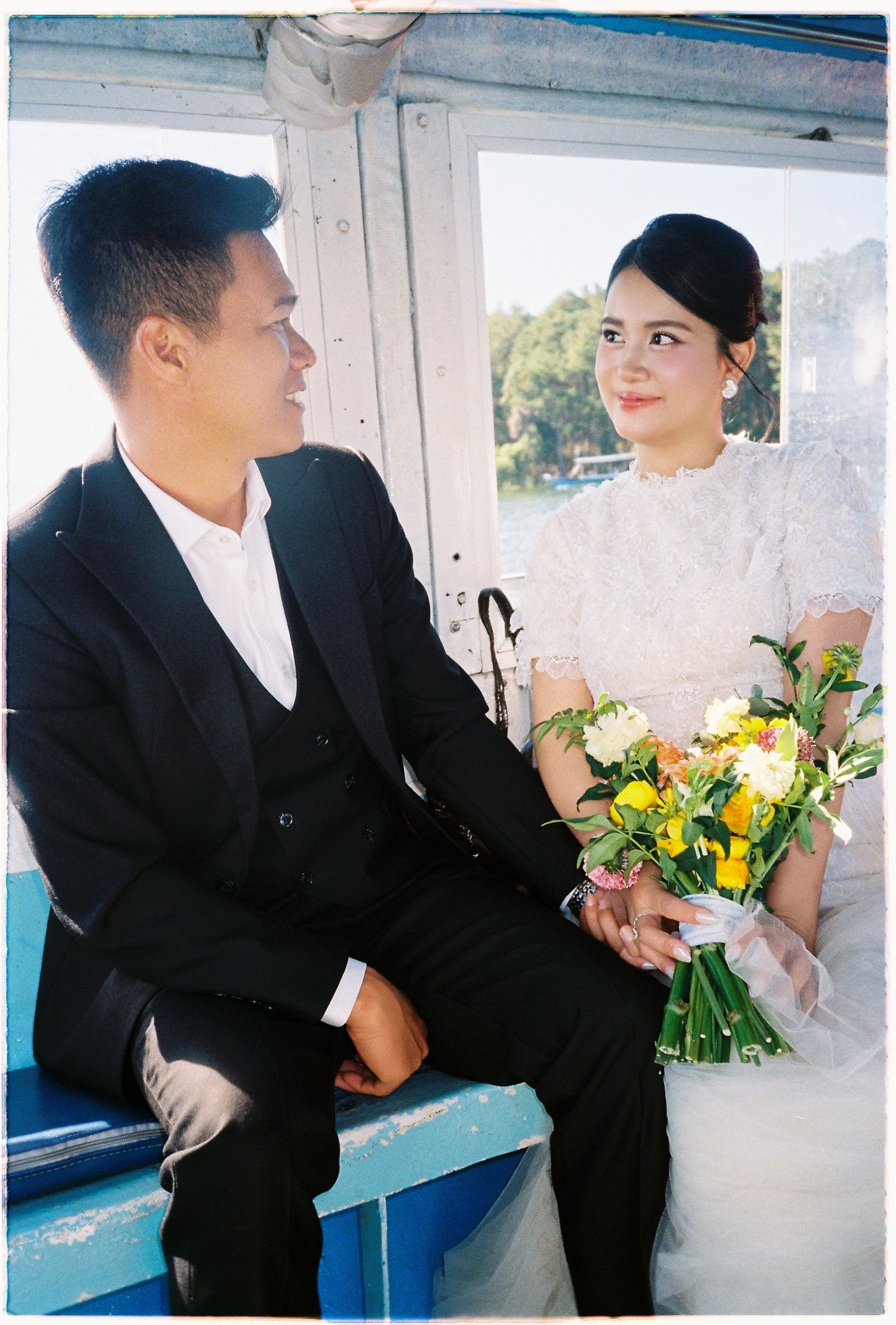 Bride and groom sit together on boat, bride holding yellow and white bouquet, smiling warmly.