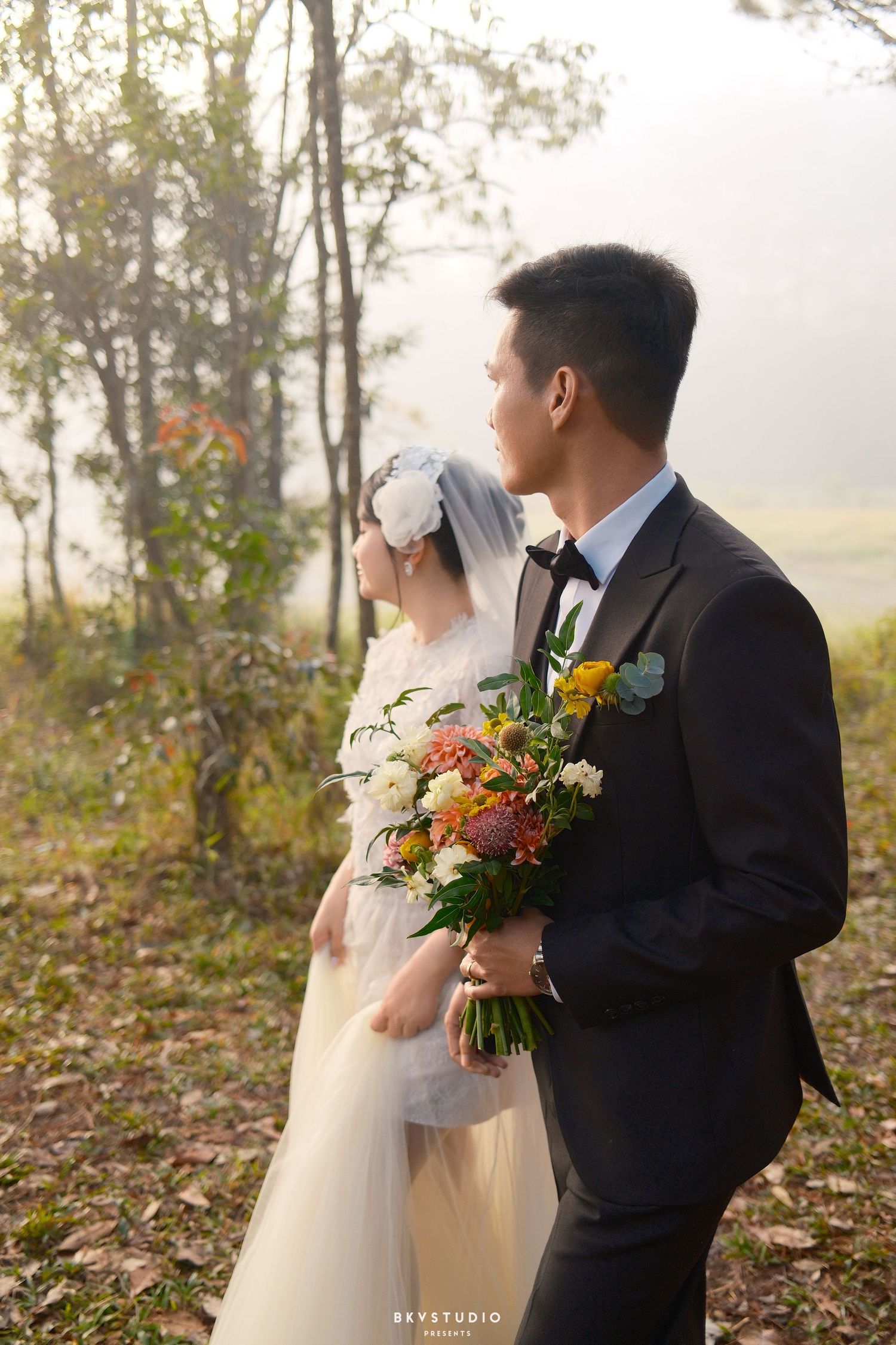 Wedding couple stands together in misty pine forest, groom holding vibrant floral bouquet in black tuxedo.