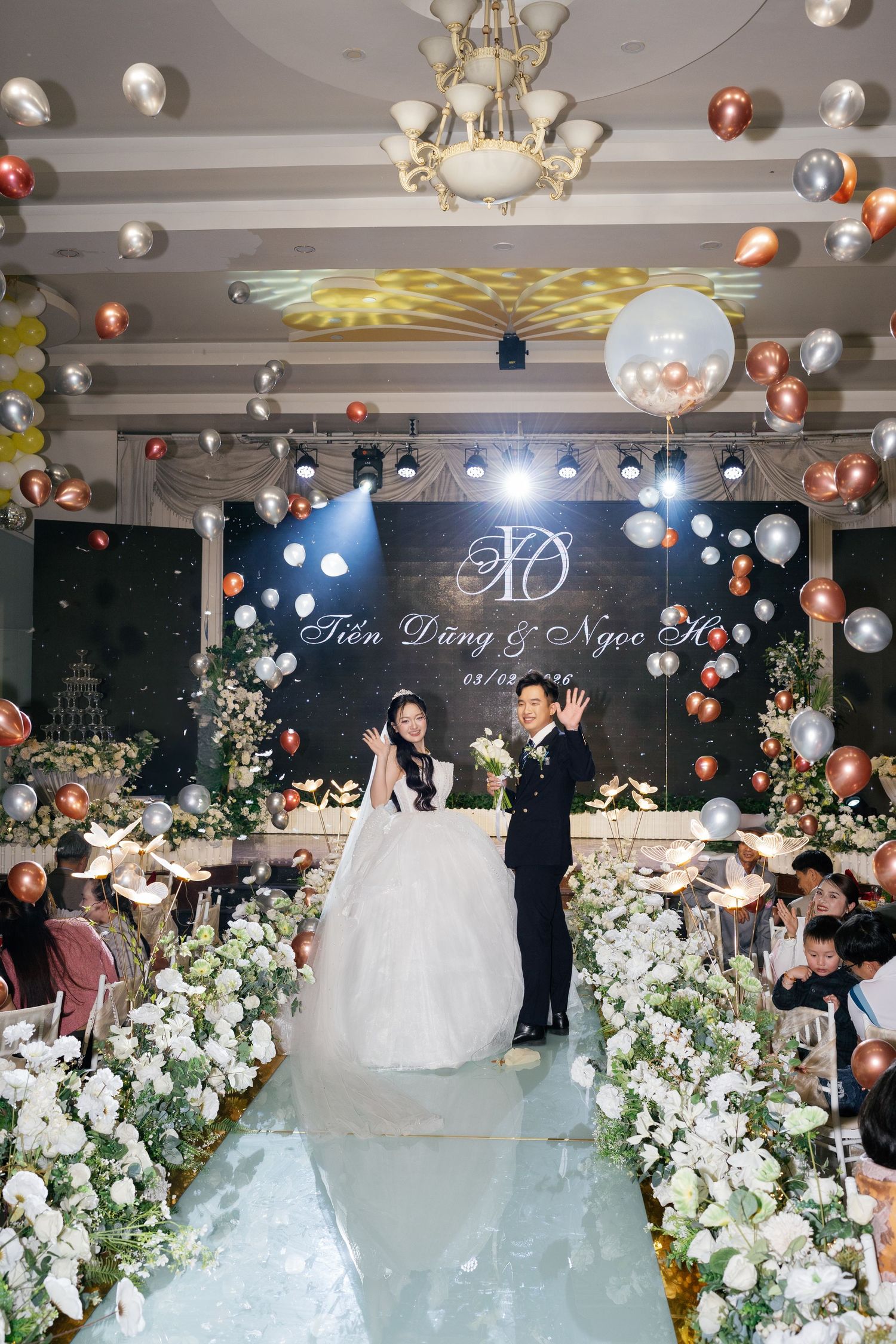 Vietnamese bride and groom celebrate on decorated wedding reception stage with falling balloons.