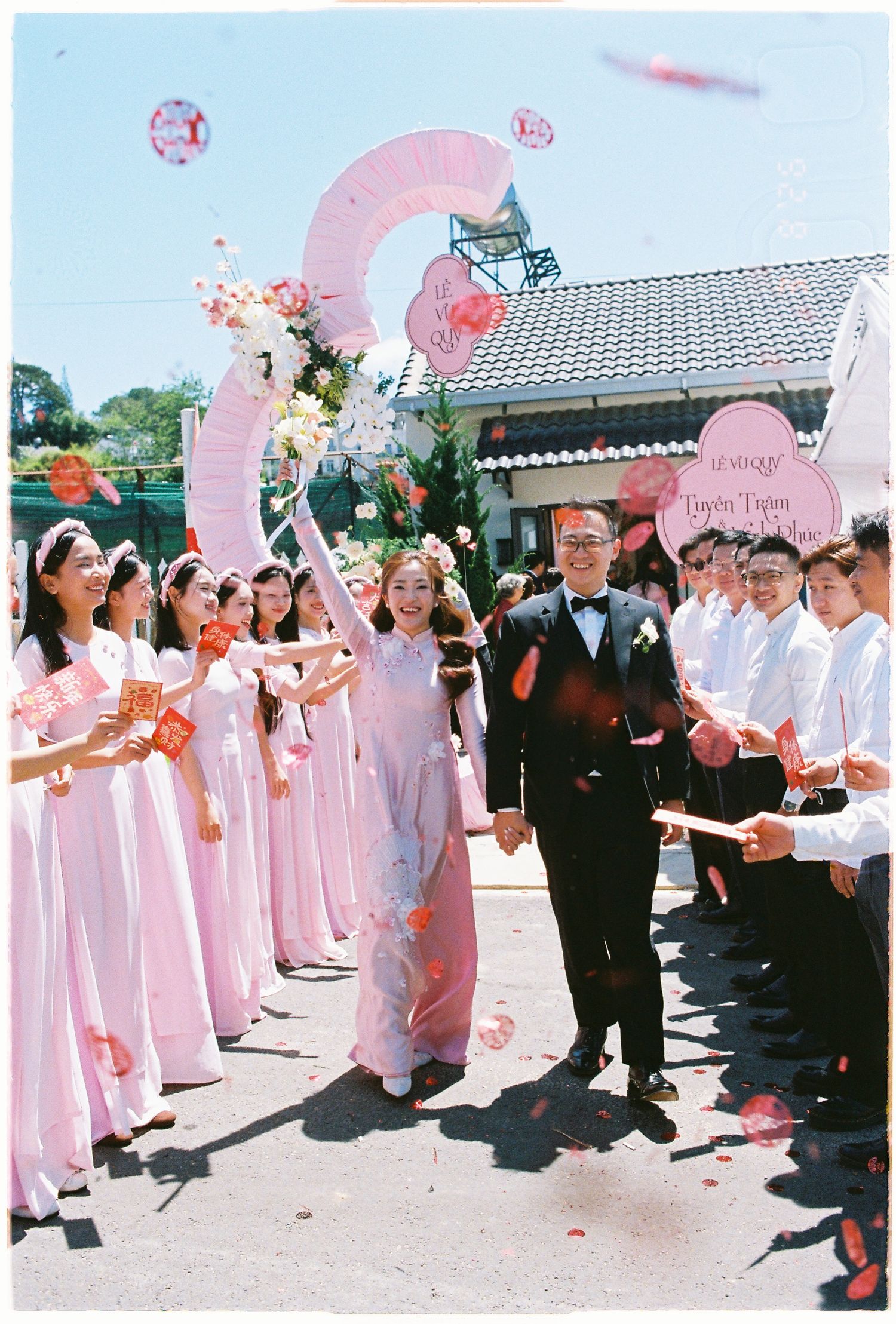 Bride and groom walking through a line of pink ao dai bridesmaids under a floral arch outdoors.