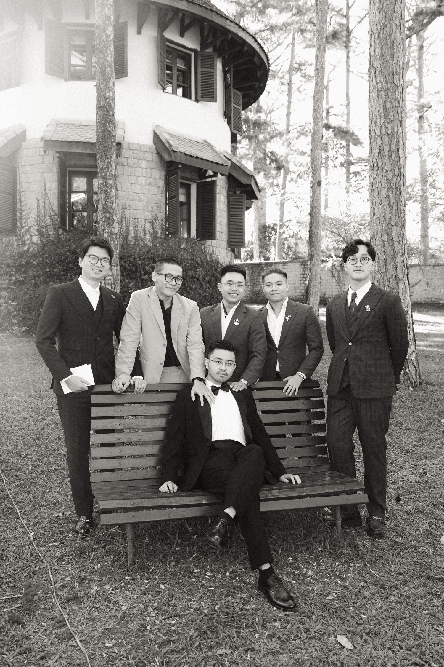 Black and white photo of groom and groomsmen posing on a bench among pine trees.