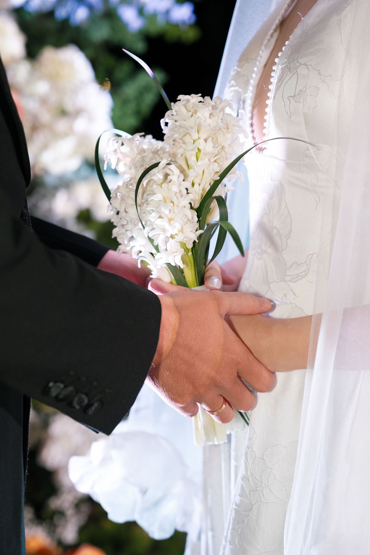 Close-up of bride and groom holding hands with white hyacinth bouquet during wedding ceremony.