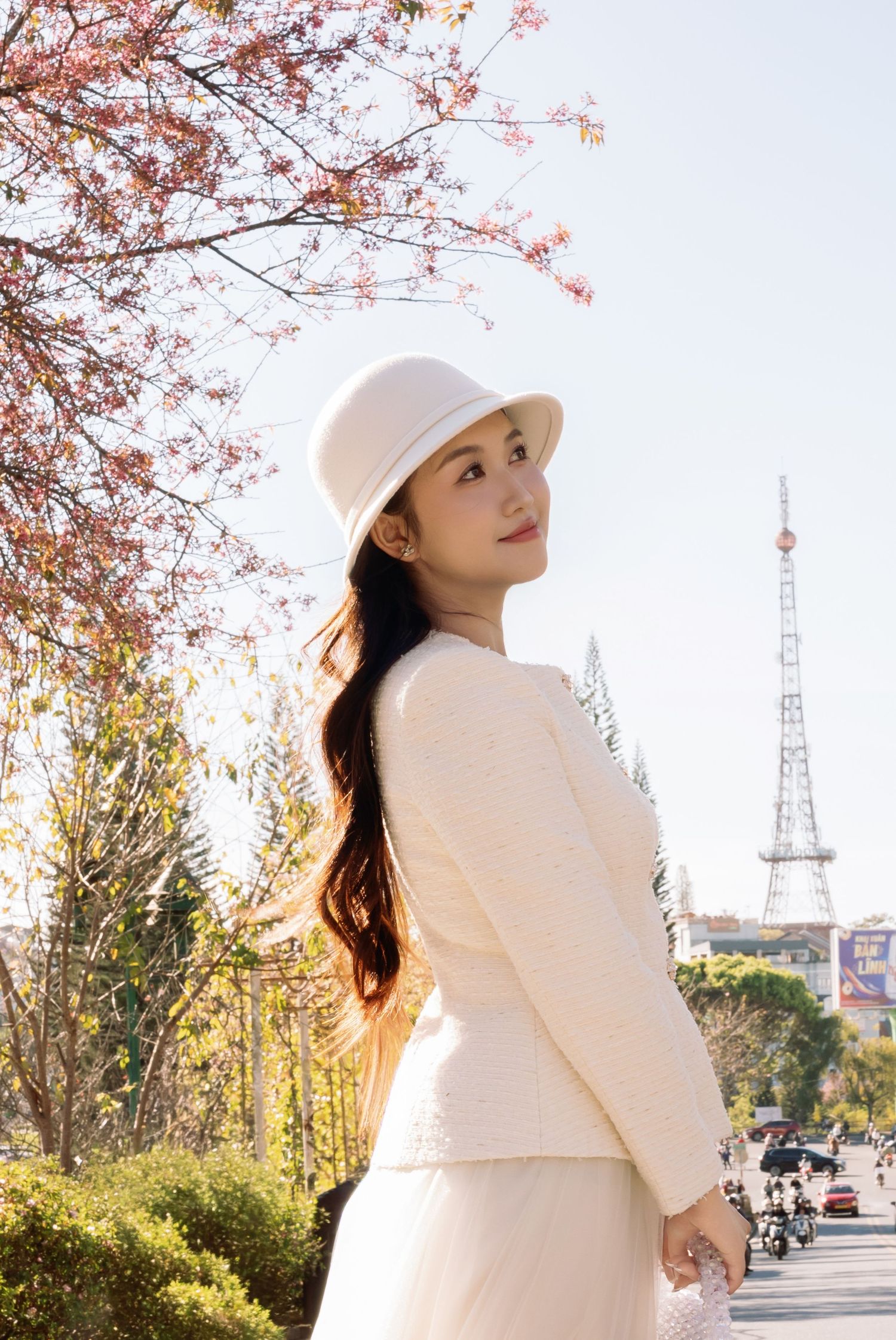 A young woman in a cream outfit and white hat poses gracefully near blooming cherry blossom trees outdoors.