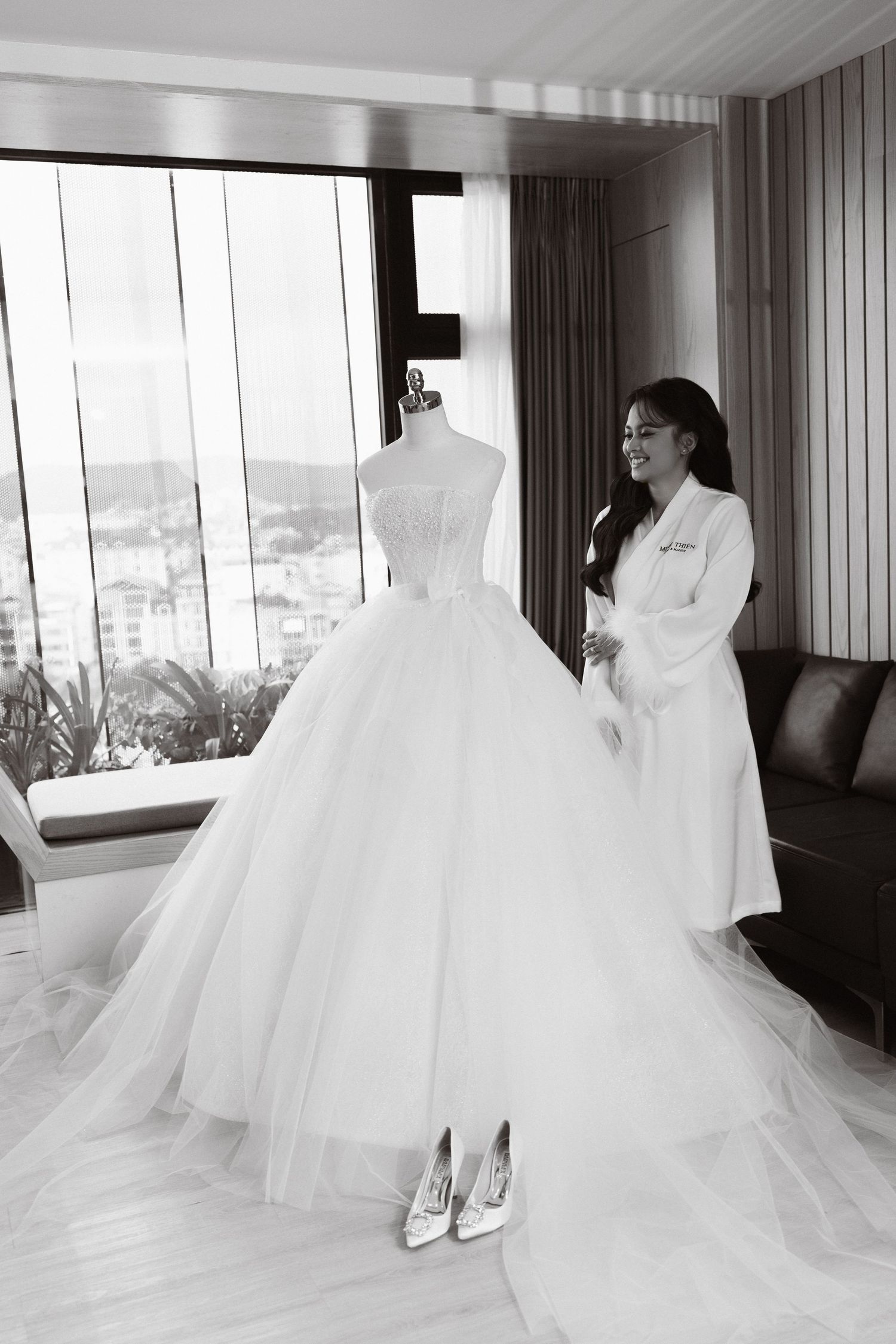 Black and white photo of a bride admiring her elegant ball gown wedding dress on a mannequin in a luxurious hotel room.