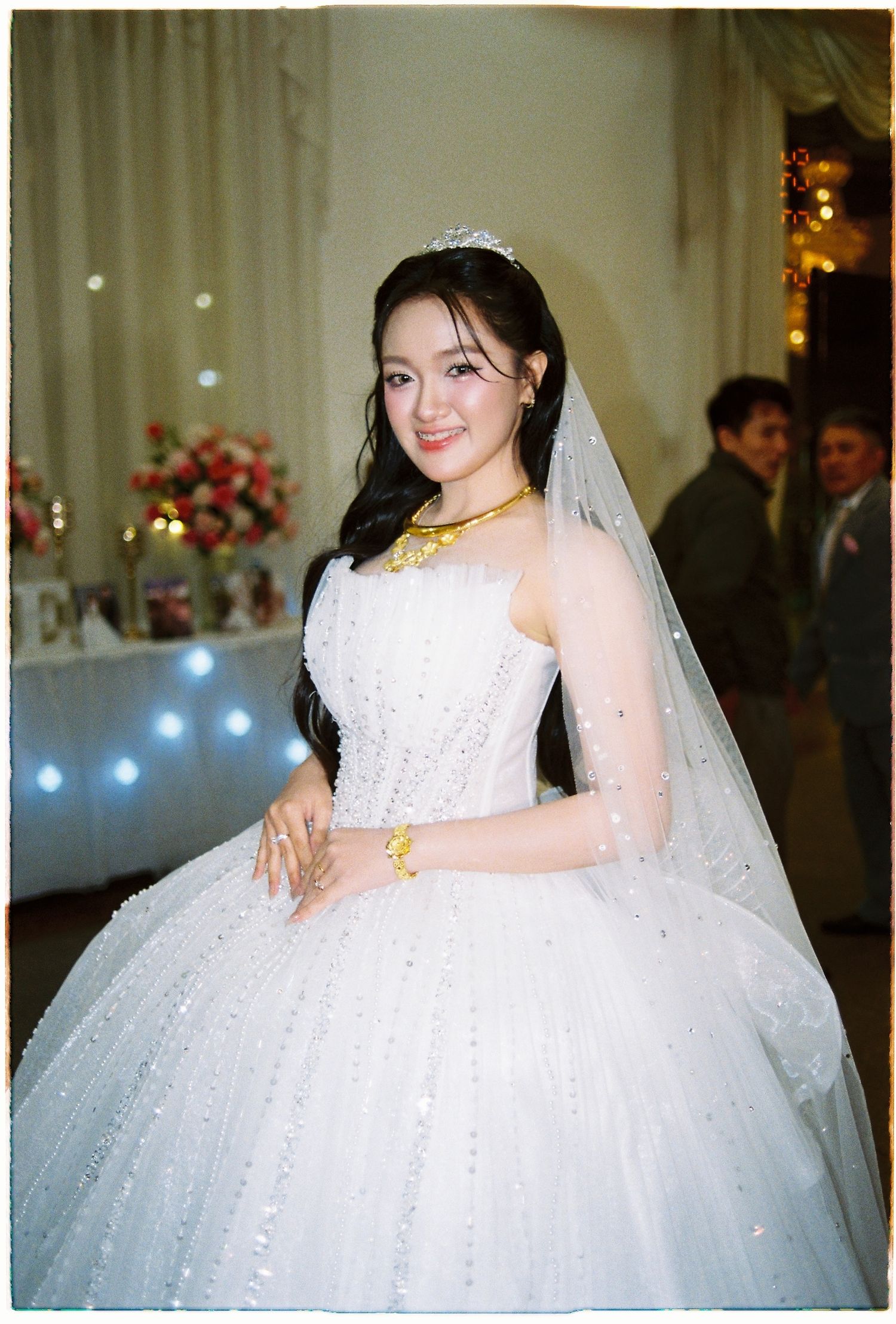 Vietnamese bride in sparkling white ballgown with tiara, veil, and gold jewelry smiling at camera.