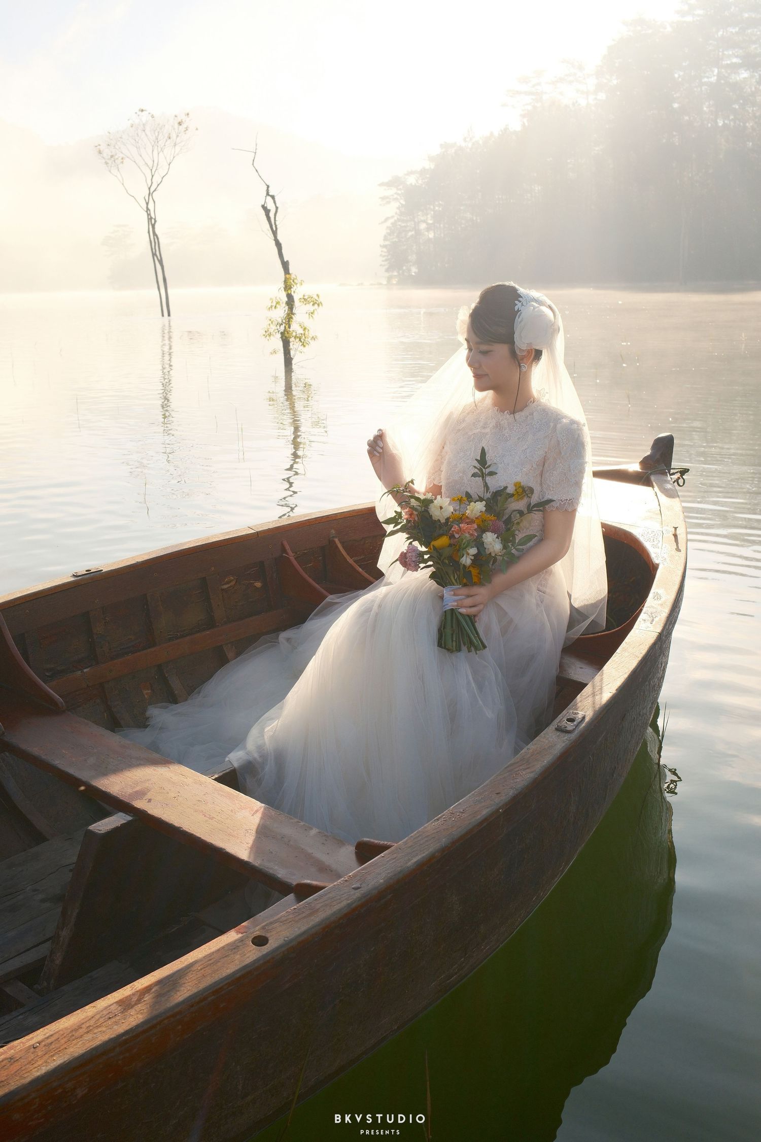 Elegant bride in white gown sitting alone in a wooden boat on a misty serene lake at sunrise.