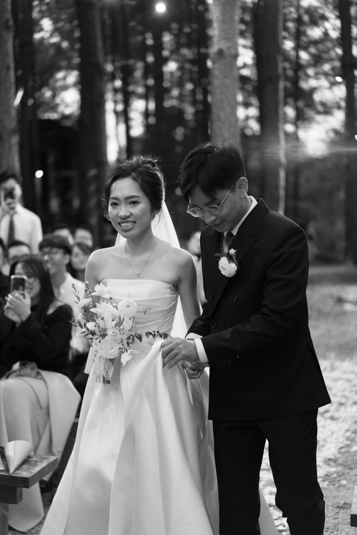 Black and white photo of smiling Asian bride holding bouquet walking down aisle with groom in suit at outdoor wedding.