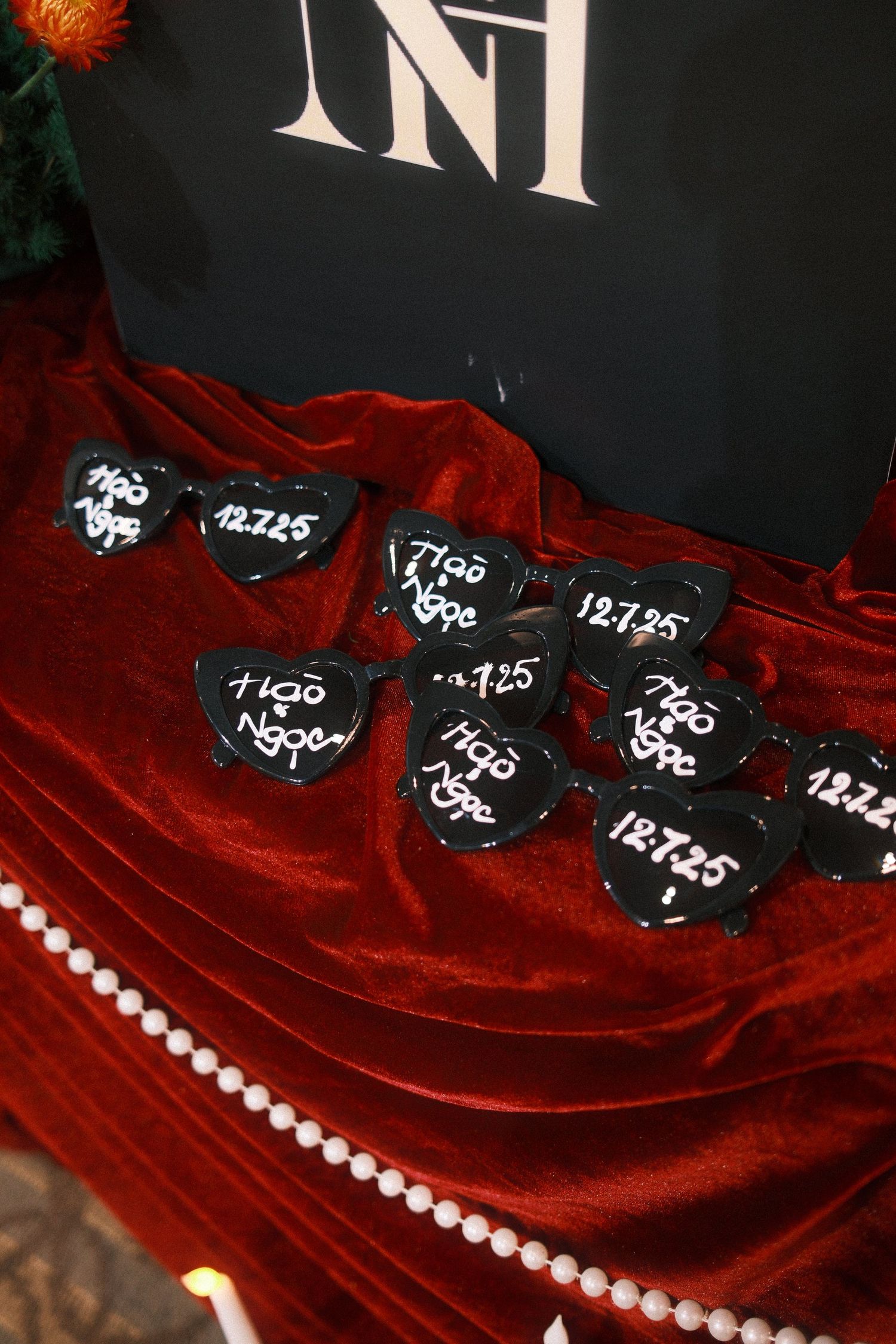 Black heart-shaped sunglasses with white script reading Ngoc 12.7.25 displayed on red velvet fabric with pearl trim.
