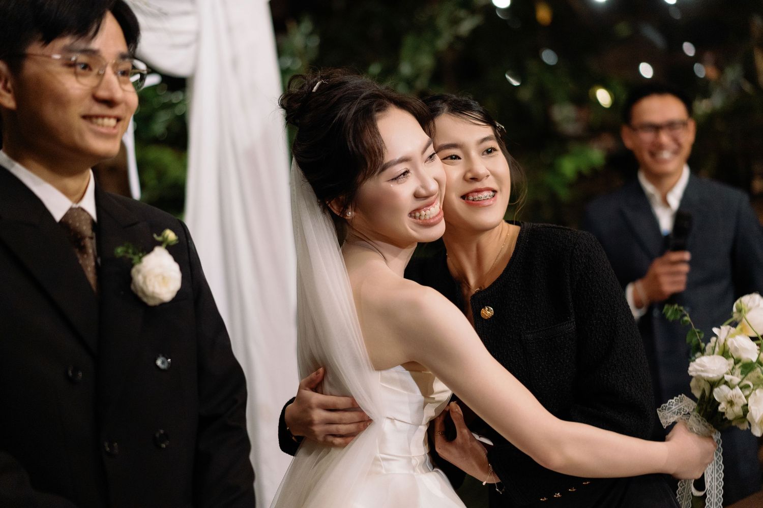 Bride in white veil shares joyful embrace with guest at romantic outdoor evening wedding.