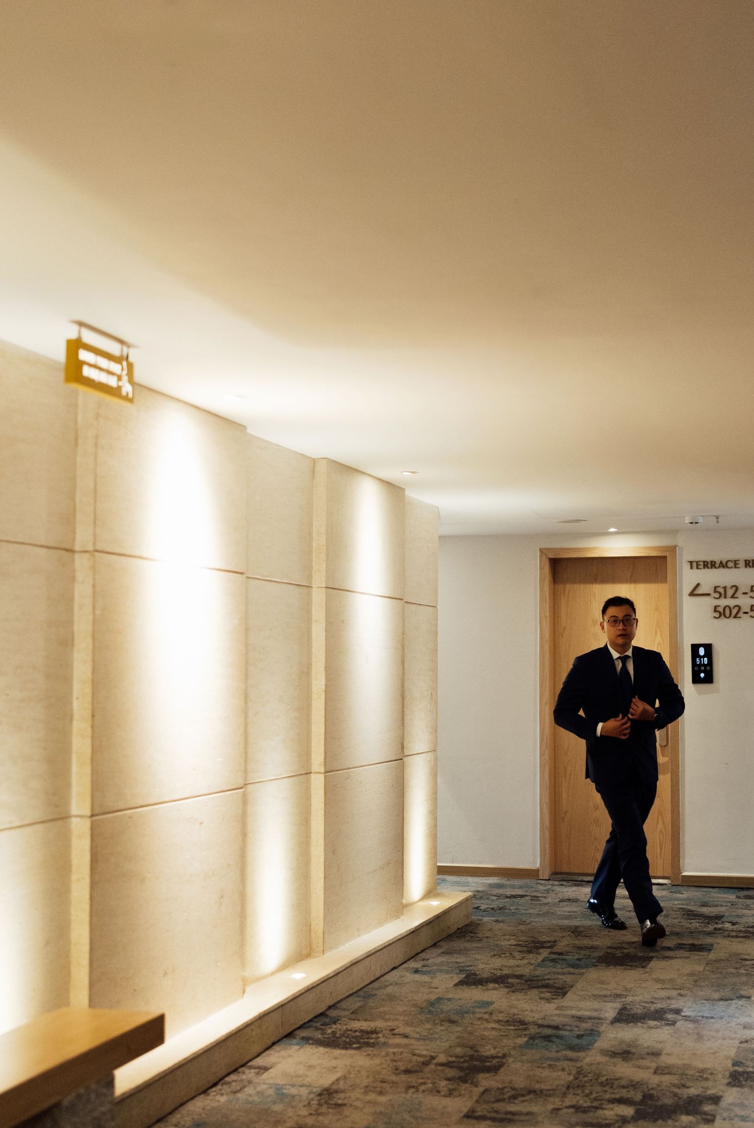 A businessman in a dark suit walks through a modern hotel corridor with warm lighting and stone walls.