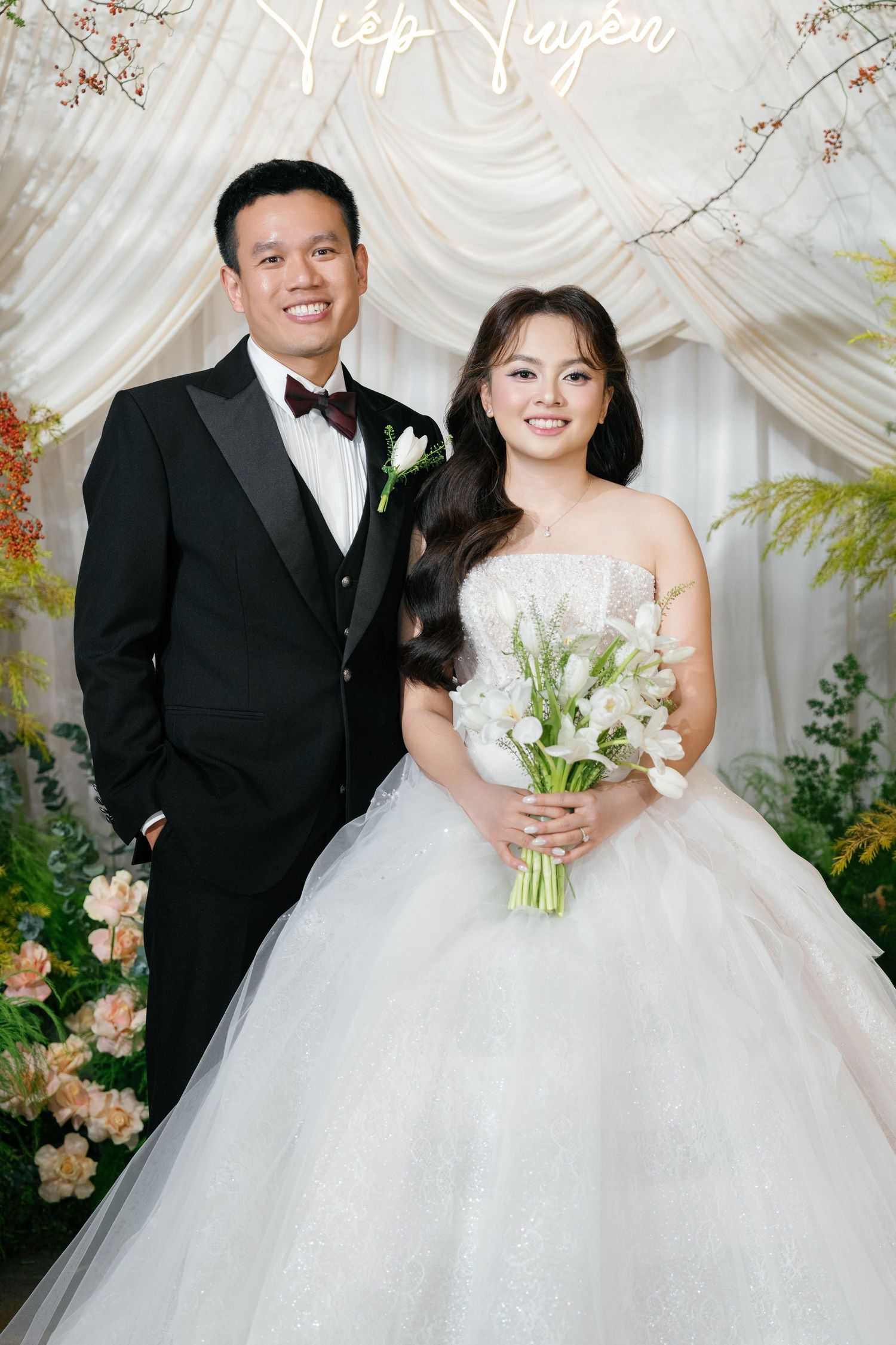 A smiling couple in wedding attire pose before a white floral backdrop at their elegant reception.