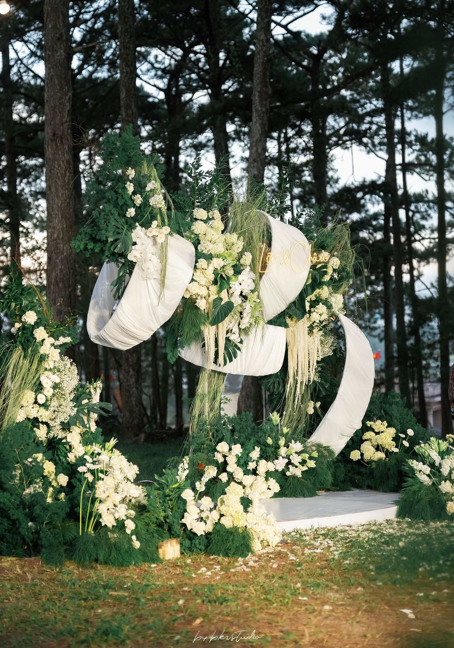 Elegant outdoor wedding arch adorned with white draping, greenery, and white floral arrangements in pine forest.