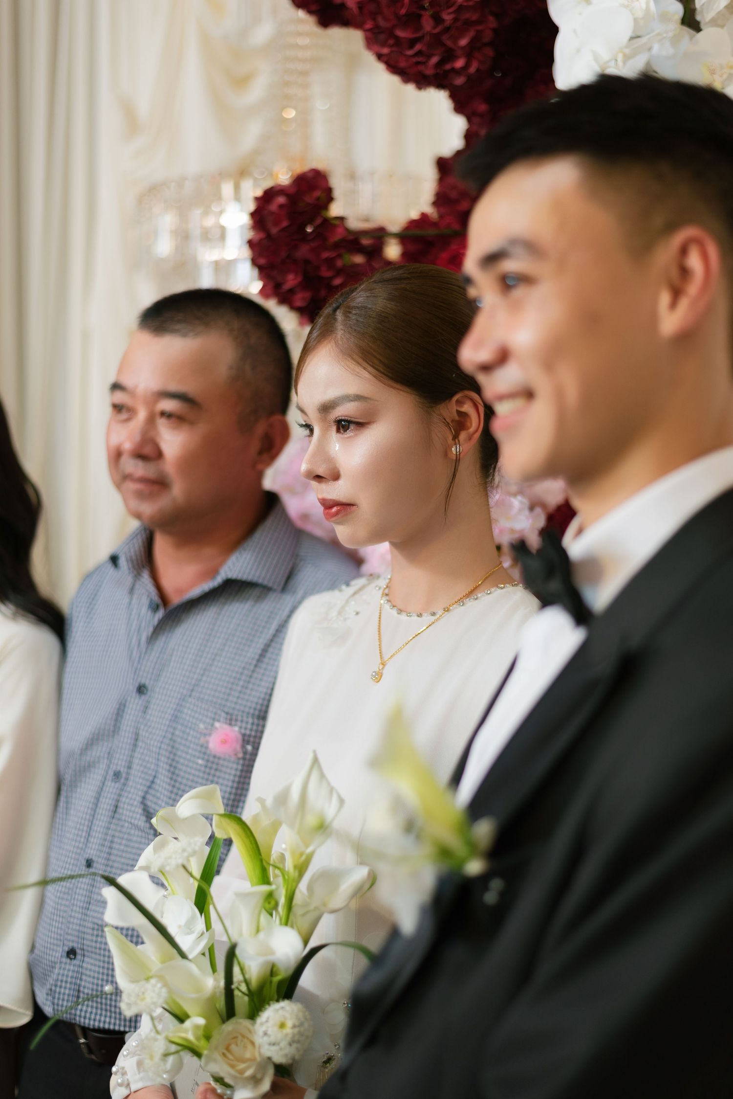 Bride holding white lilies stands between two men at an elegant wedding ceremony with floral decorations.