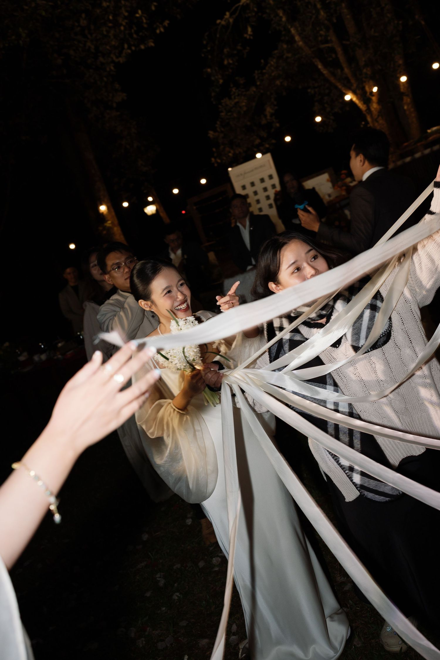 Guests celebrate at an outdoor wedding reception at night, playing with white ribbons under string lights.