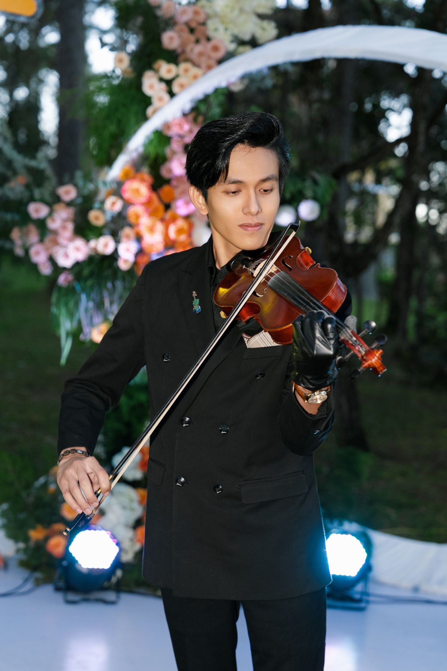 Musician in black suit playing violin at outdoor wedding ceremony with floral arch and stage lights.