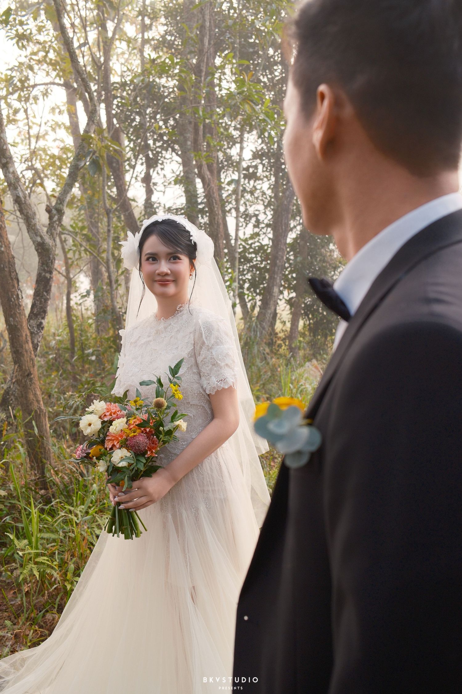 Bride in lace wedding dress holding colorful bouquet smiles at groom in black suit during outdoor forest wedding ceremony.
