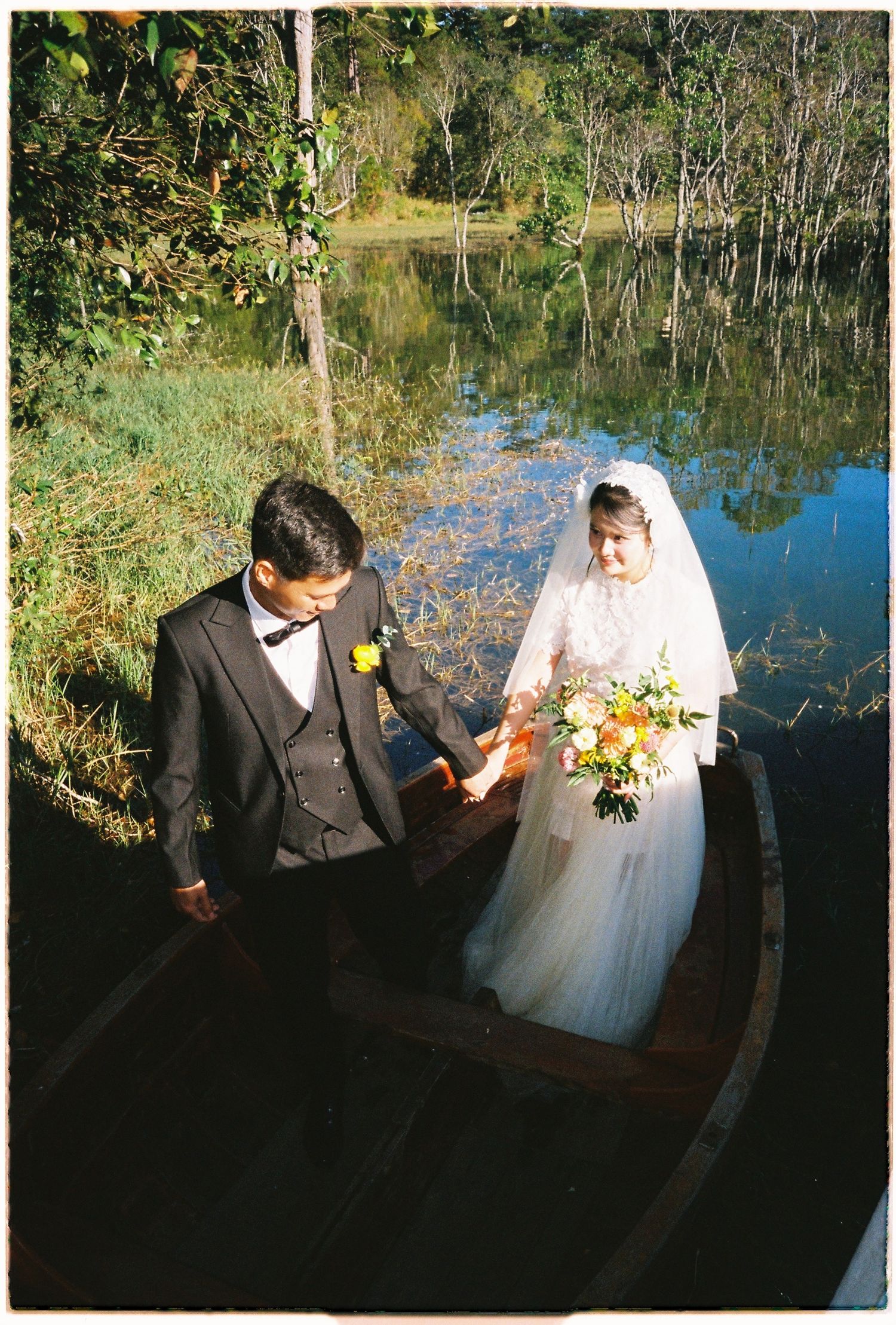 Bride and groom holding hands stepping into wooden rowboat by reflective lake in lush forest setting.