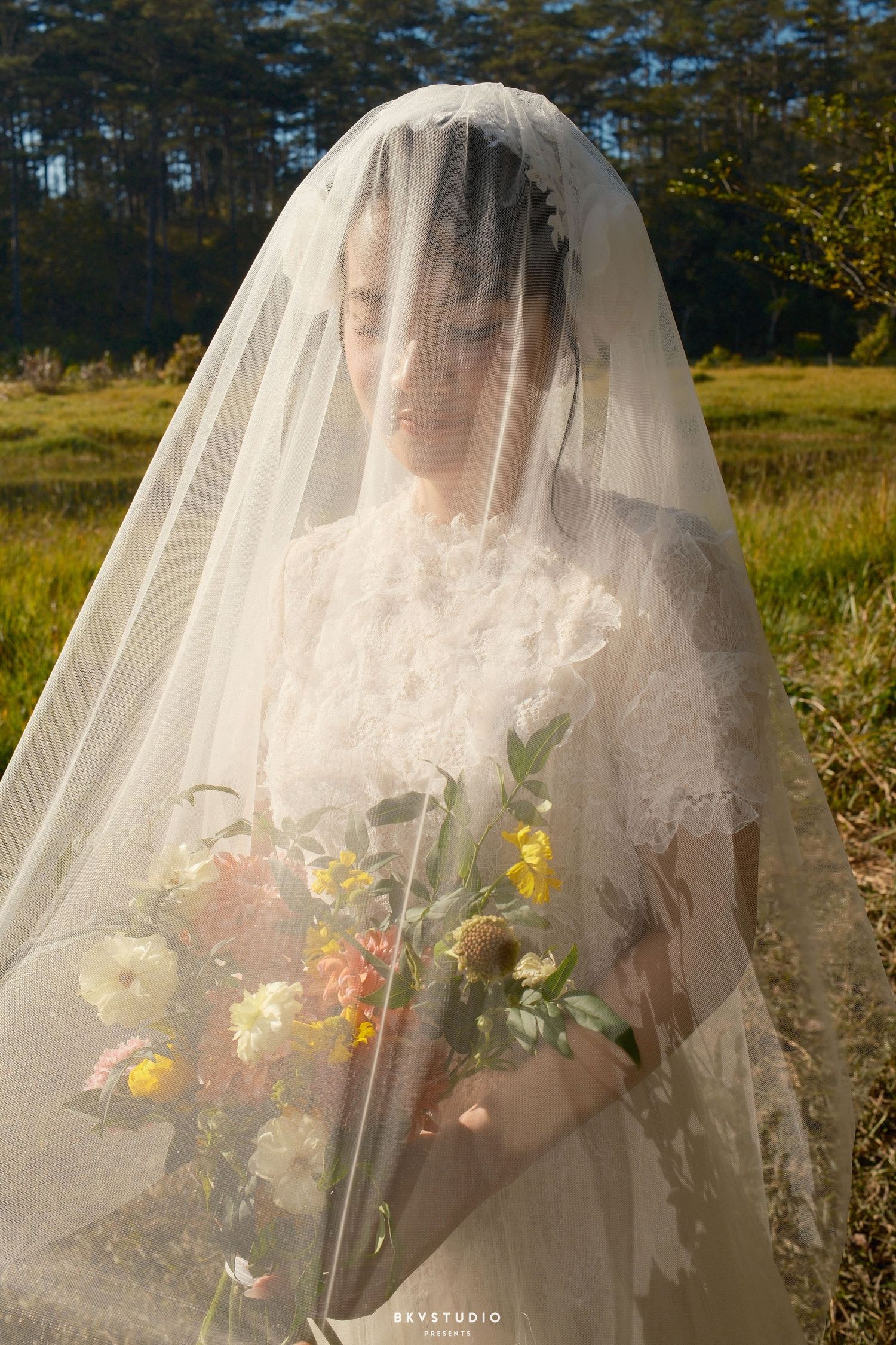 Bride covered by flowing veil holds colorful bouquet while standing in sunlit outdoor meadow.