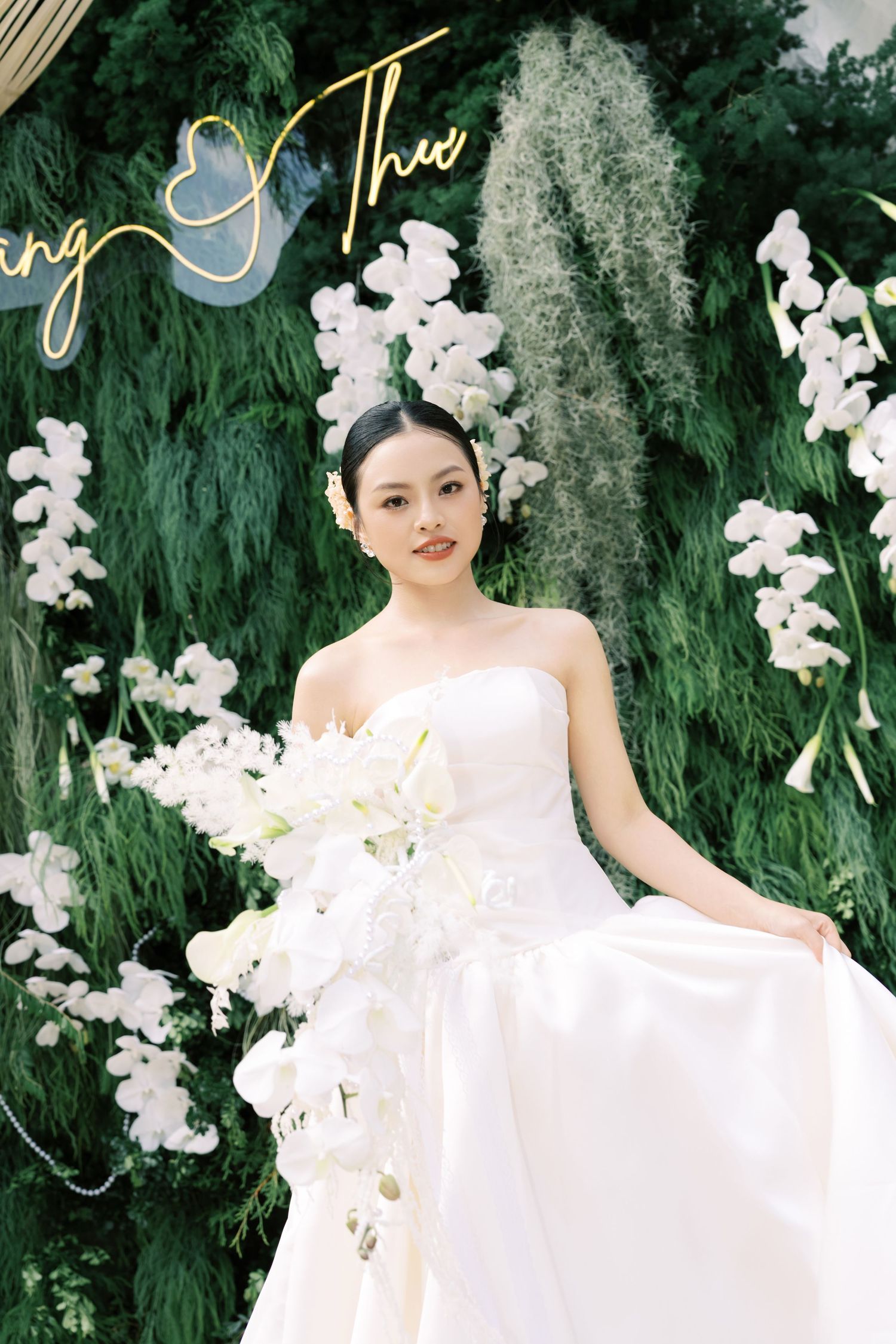 Bride in strapless white gown holding cascading white bouquet in front of lush green floral backdrop.