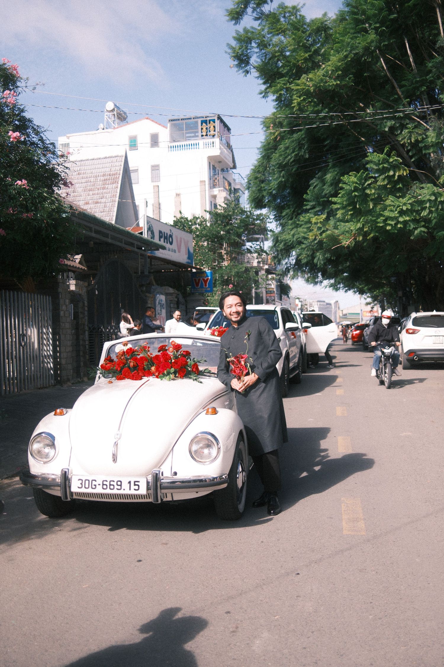 Woman in gray dress poses beside a white VW Beetle decorated with red roses on a tree-lined street.